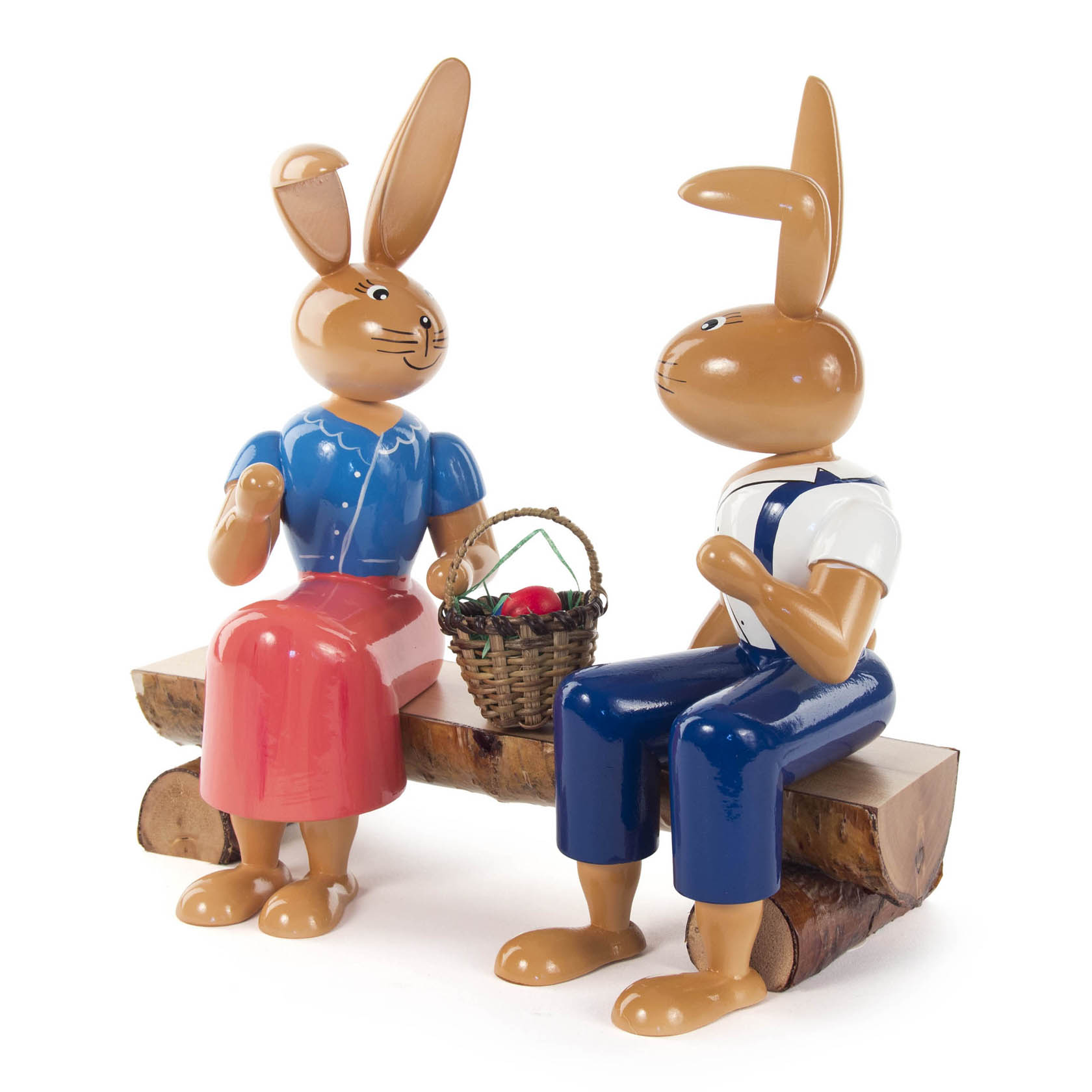 Two wooden bunny figurines sitting on a bench. The left bunny wears a blue blouse and red skirt, holding a basket of eggs. The right bunny wears blue overalls.