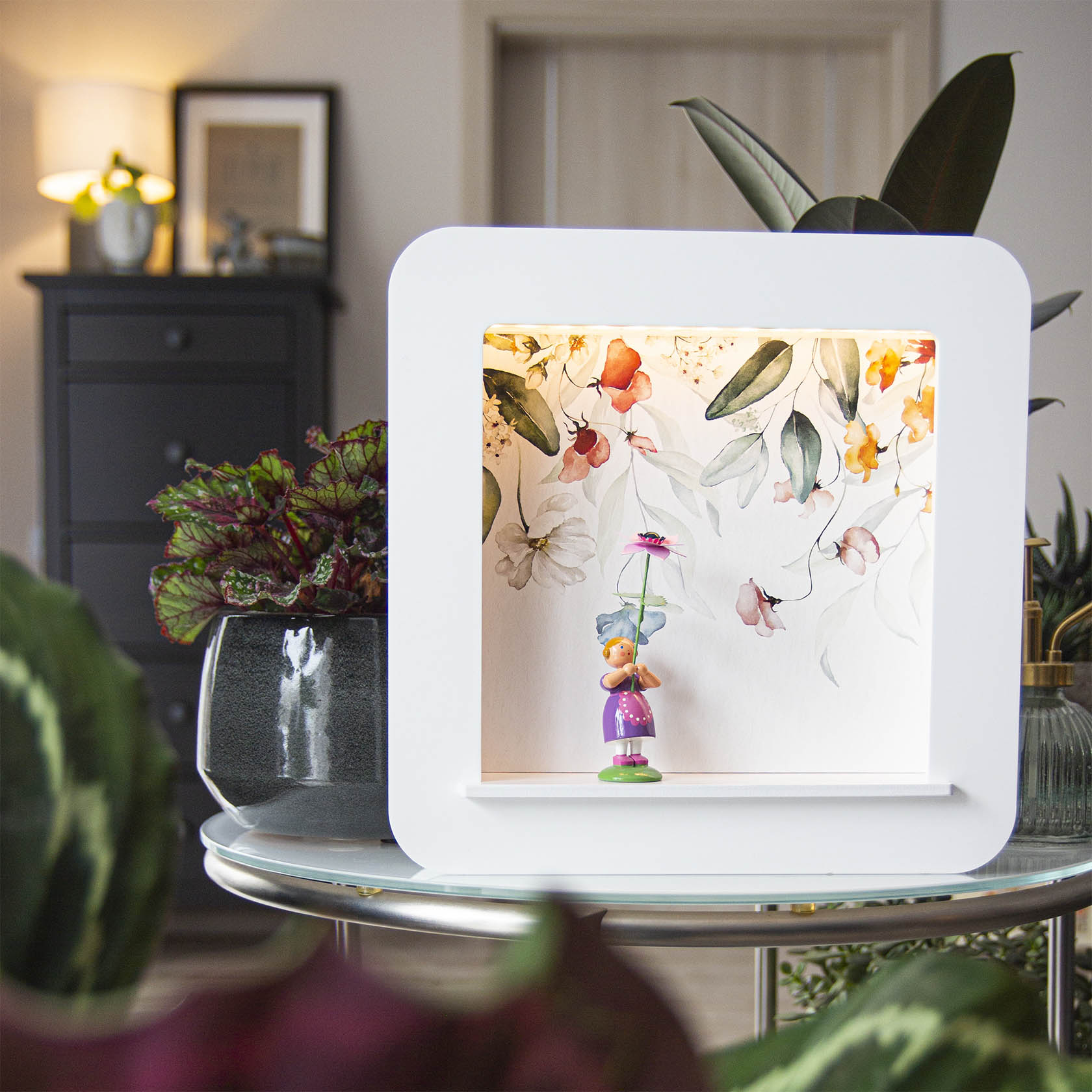 Weißer Objektrahmen mit LED-Beleuchtung, in dem eine kleine, bunte Figur mit einem Blumenmotiv im Hintergrund steht. Pflanzen umgeben den Rahmen.