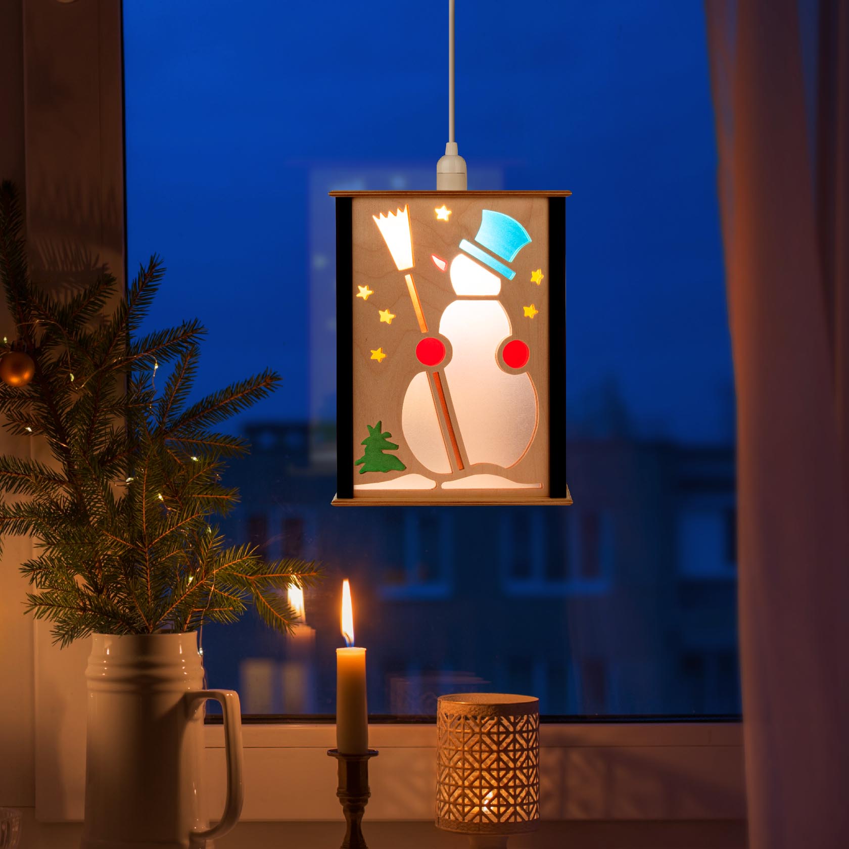 A glowing square lantern in the window with a snowman design, featuring stars and a broom, with a candle and a small fir tree beside it.