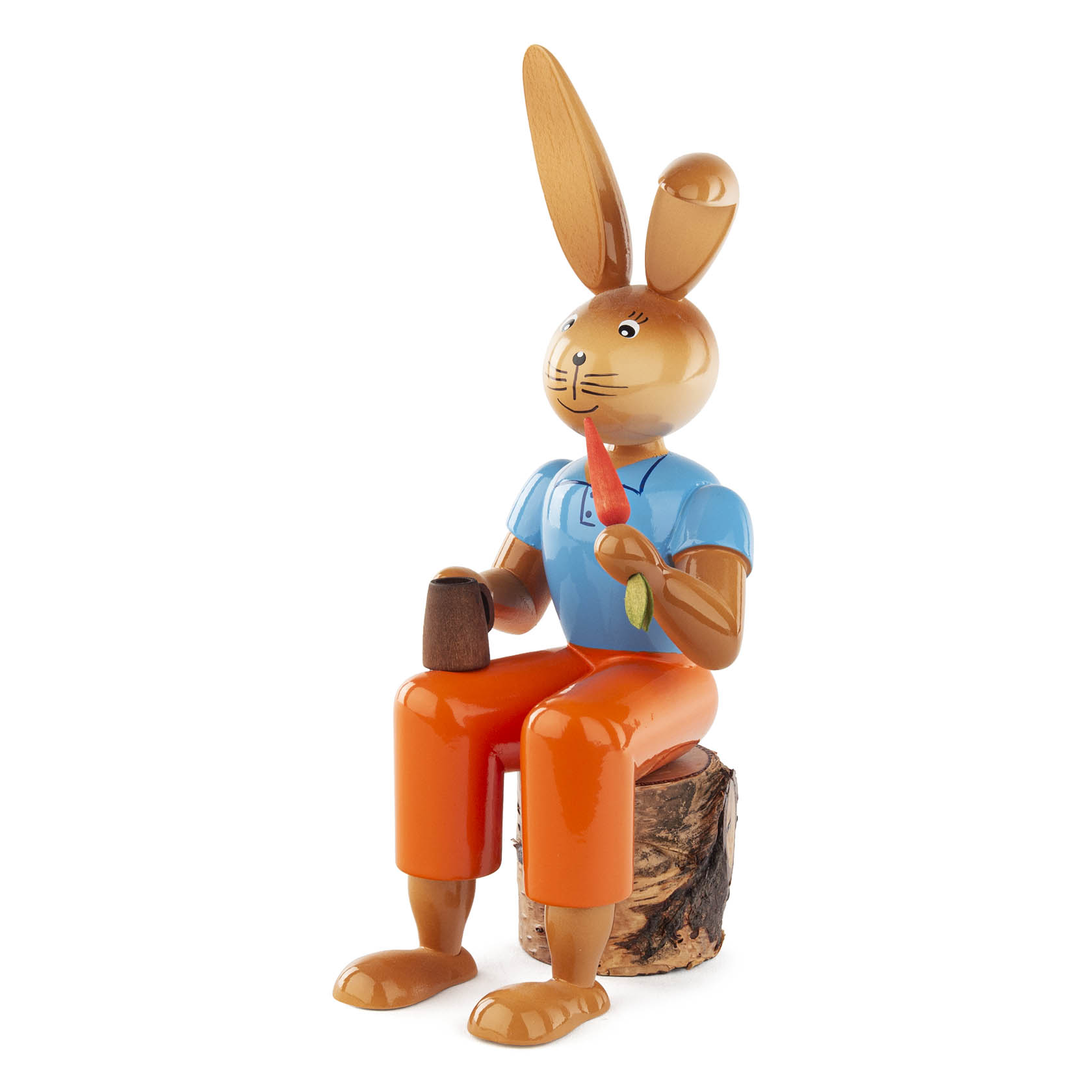 Holzhase in Blau und Orange sitzt auf einem Baumstumpf und hält eine Karotte in der rechten Hand.
