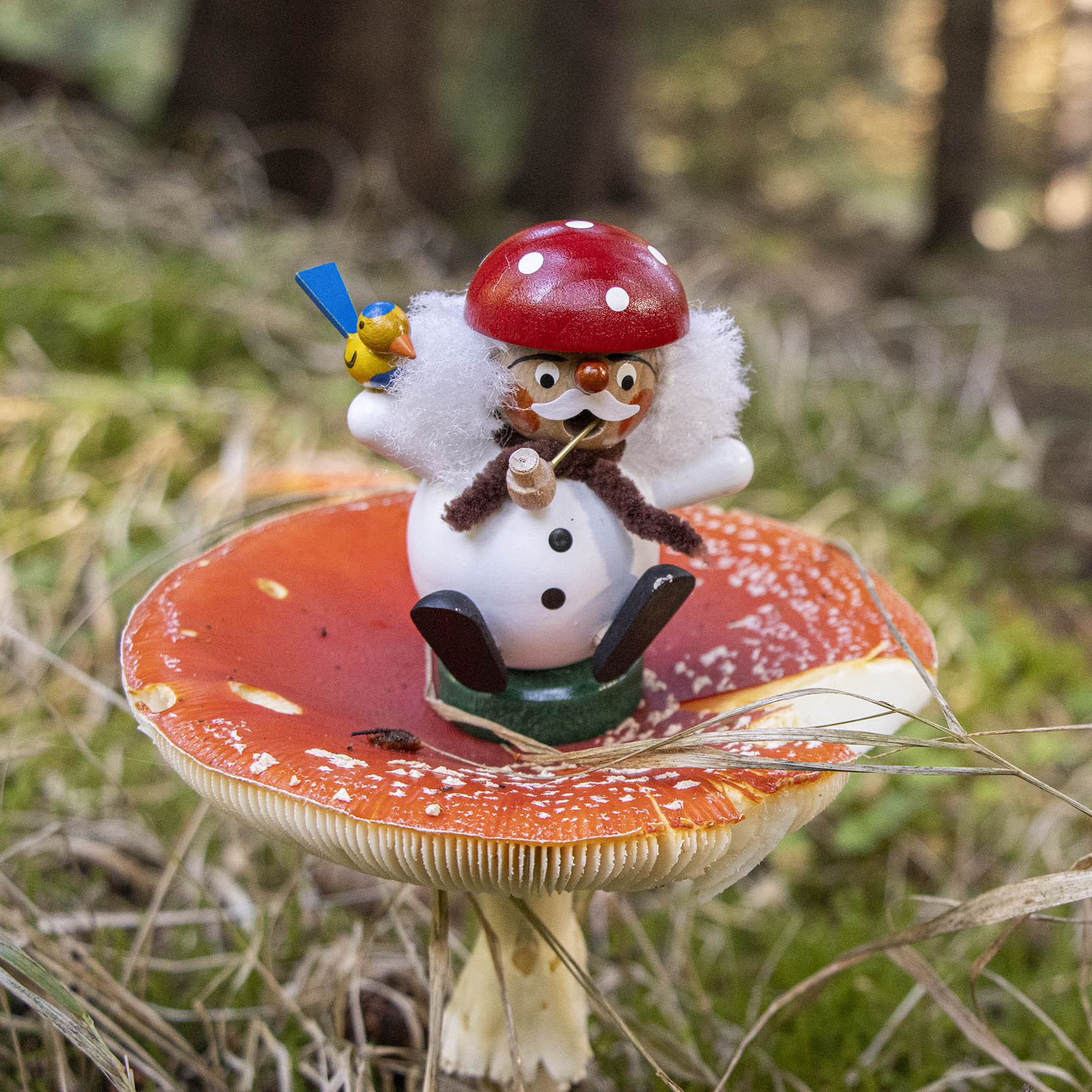 Ein kugelförmiger Räuchermann mit rotem Pilzhut sitzt auf einem Fliegenpilz. Er hat einen weißen Bart, eine Pfeife im Mund und hält einen blauen Besen.