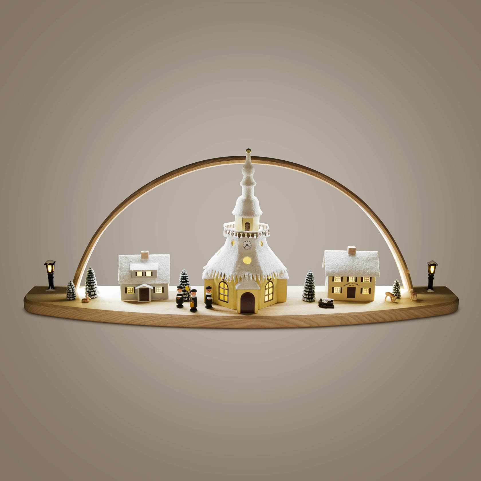 Ein Schwibbogen mit LED-Beleuchtung zeigt ein Seiffener Dorf. In der Mitte ist eine Kirche, flankiert von zwei Häusern. Kleine Bäume und Figuren stehen dekorativ daneben.