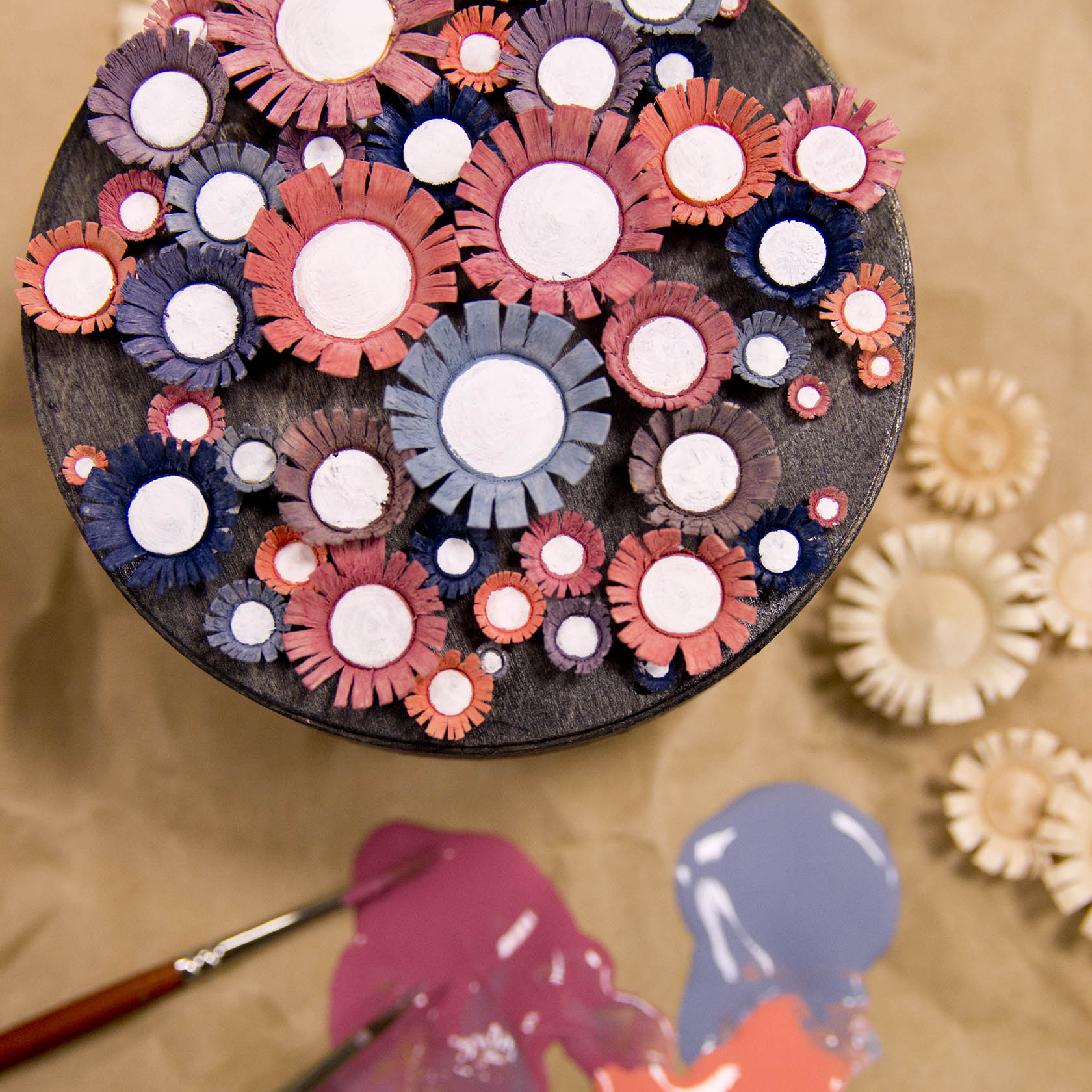 Collection of small handmade clay flower sculptures in pink, purple, and blue on a round black stand, with paintbrushes nearby.