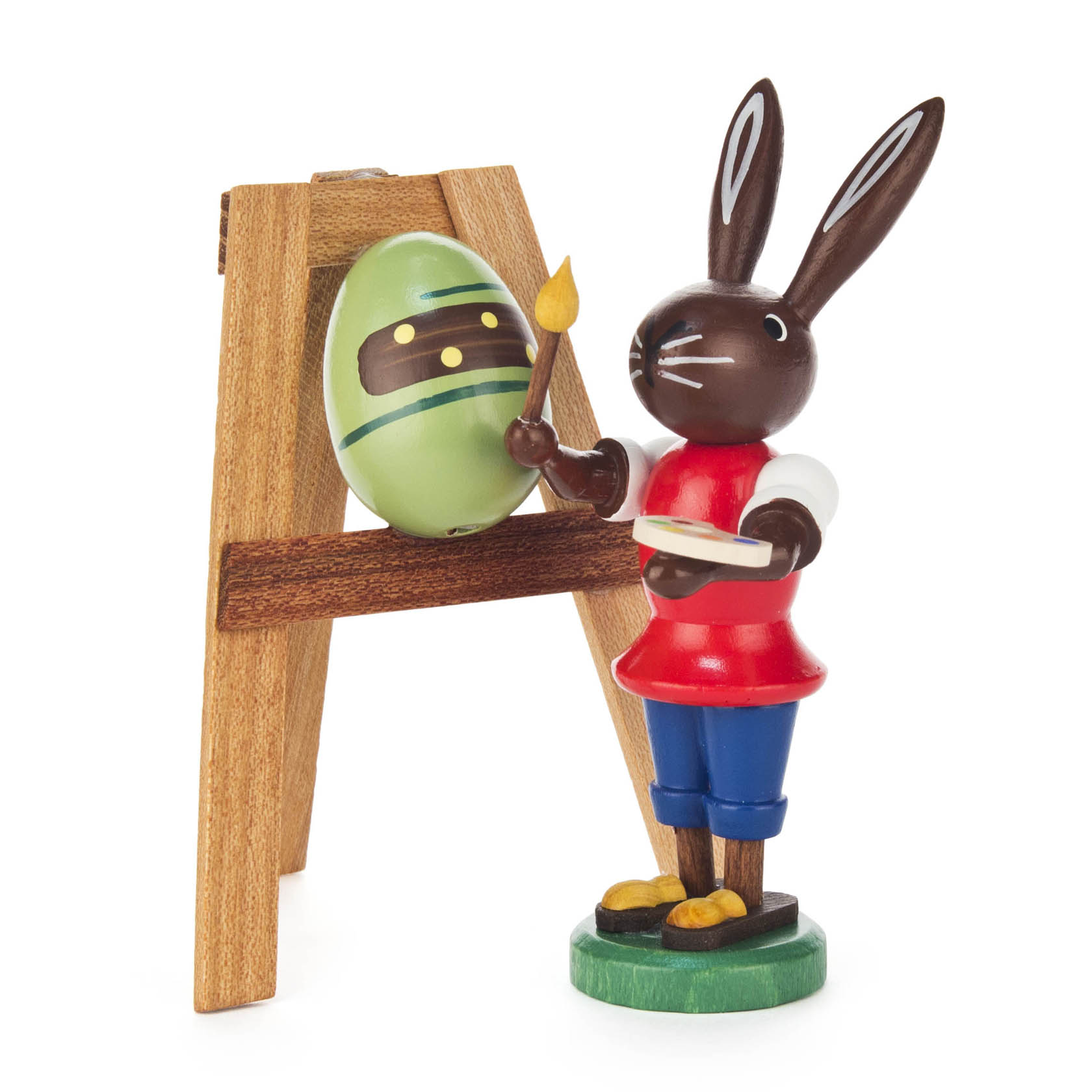 Holzfigur eines Hasen in rotem Shirt und blauer Hose, der mit einem Pinsel vor einer Staffelei mit einem dekorierten Ei steht. im Dregeno Online Shop günstig kaufen