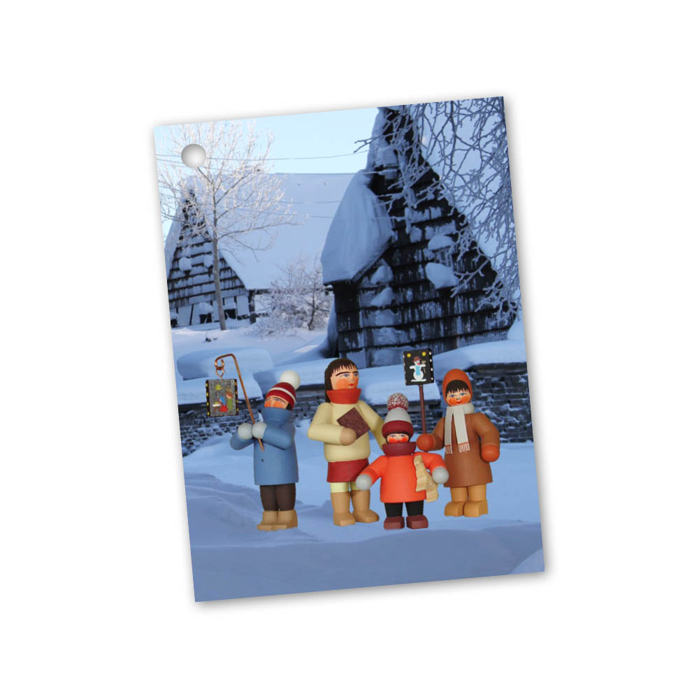 Four figurines dressed in winter clothes holding lanterns, standing in front of snow-covered chalet-style houses. im Dregeno Online Shop günstig kaufen