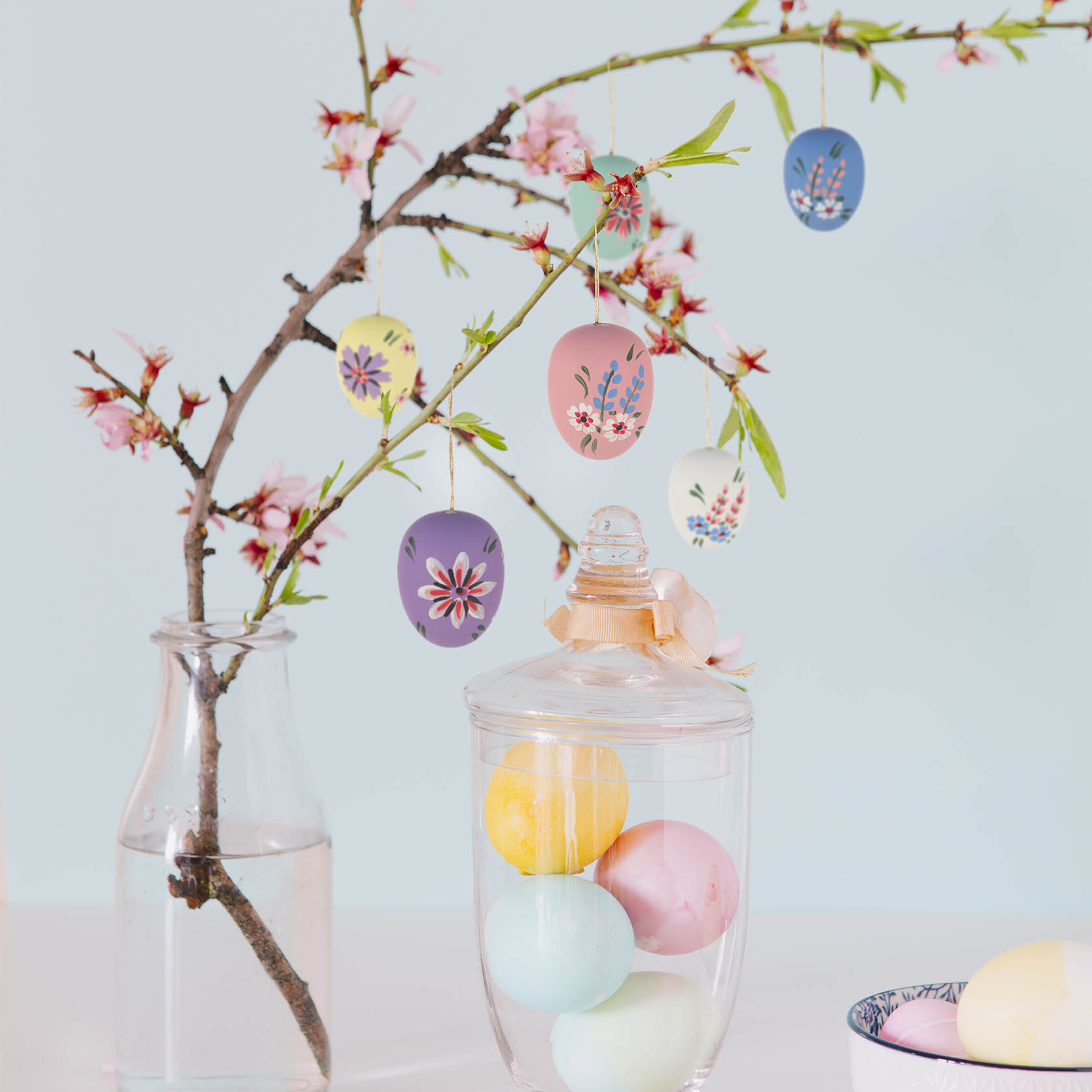 Zweig mit rosa Blüten, behangen mit sechs seidenmatten Ostereiern in pastelligen Farben und floralen Mustern. Ein Glasbehälter mit farbigen Eiern daneben.