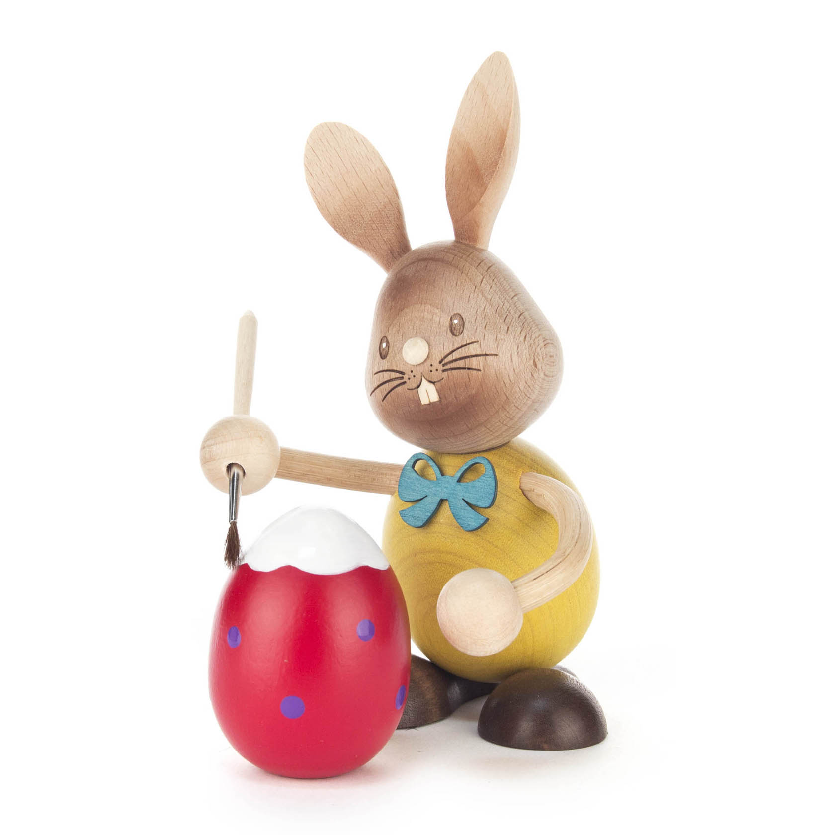 Holzhase 'Stupsi' bemalt ein rotes Osterei mit blauen Punkten und weißem oberen Rand. Er hält einen Pinsel in der rechten Pfote und trägt eine gelbe Weste mit grüner Schleife. im Dregeno Online Shop günstig kaufen