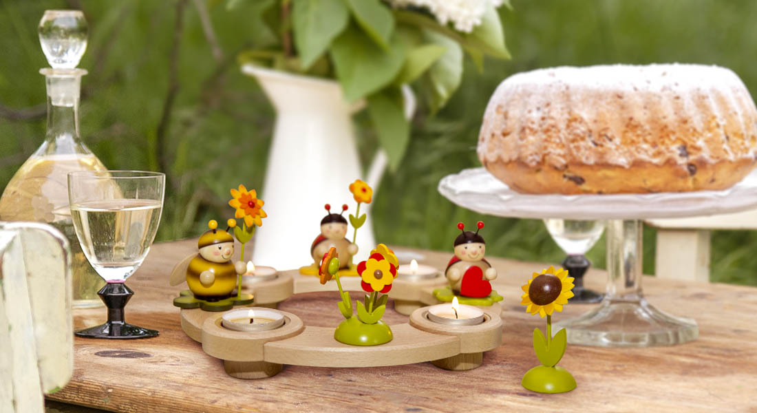 Ein runder Teelichhalter mit Käfer- und Blumenfiguren steht auf einem gedeckten Tisch im Freien