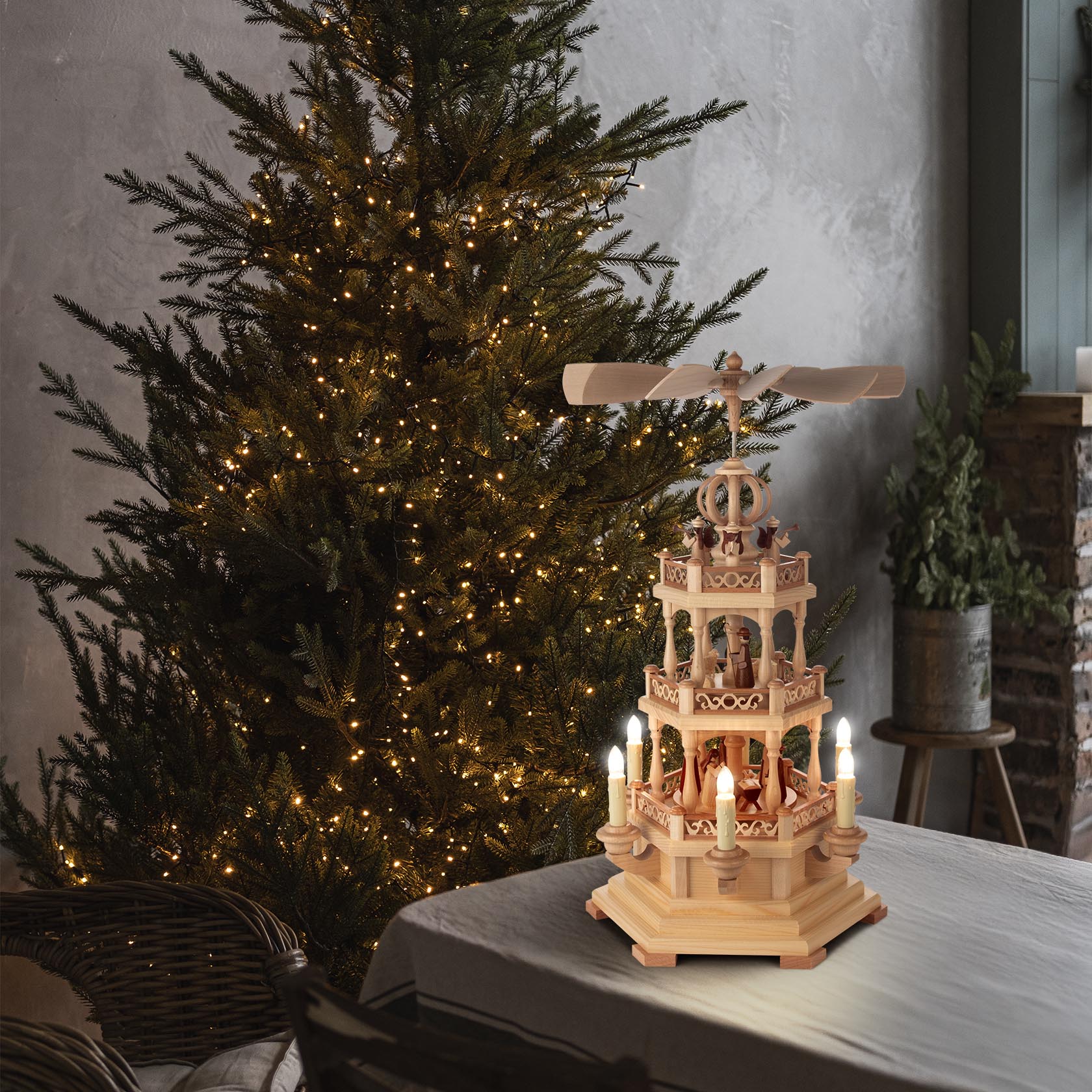 Zweistufige Weihnachts-Pyramide aus Holz mit Szenen der Christi Geburt, elektrischer Beleuchtung und elektrischem Antrieb, auf einem Tisch vor einem beleuchteten Weihnachtsbaum.