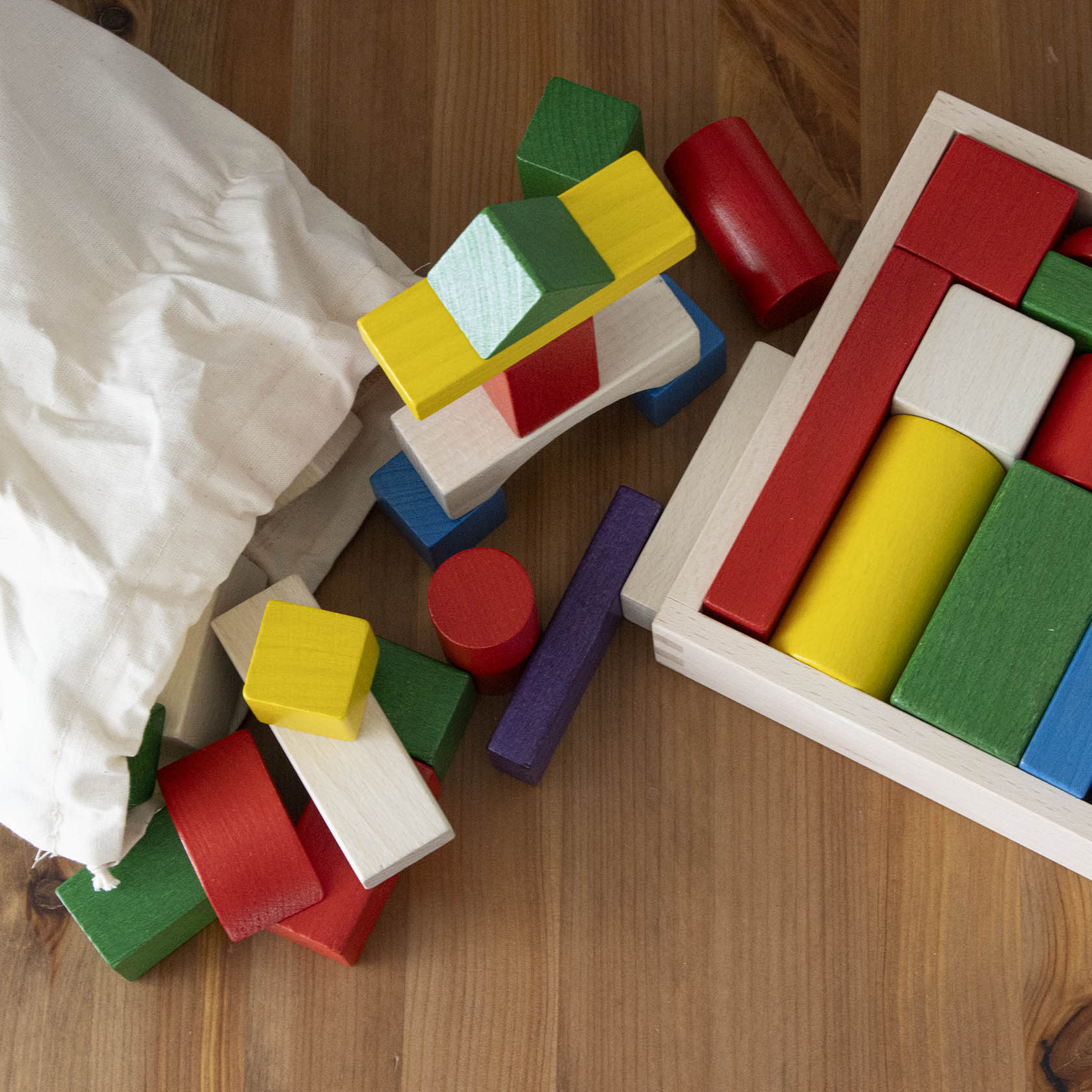 Ein gefüllter Stoffbeutel mit bunt bemalten Holzbausteinen, die auf einem Holztisch verstreut sind. Rechts daneben ein Holzrahmen mit geordneten Bausteinen.