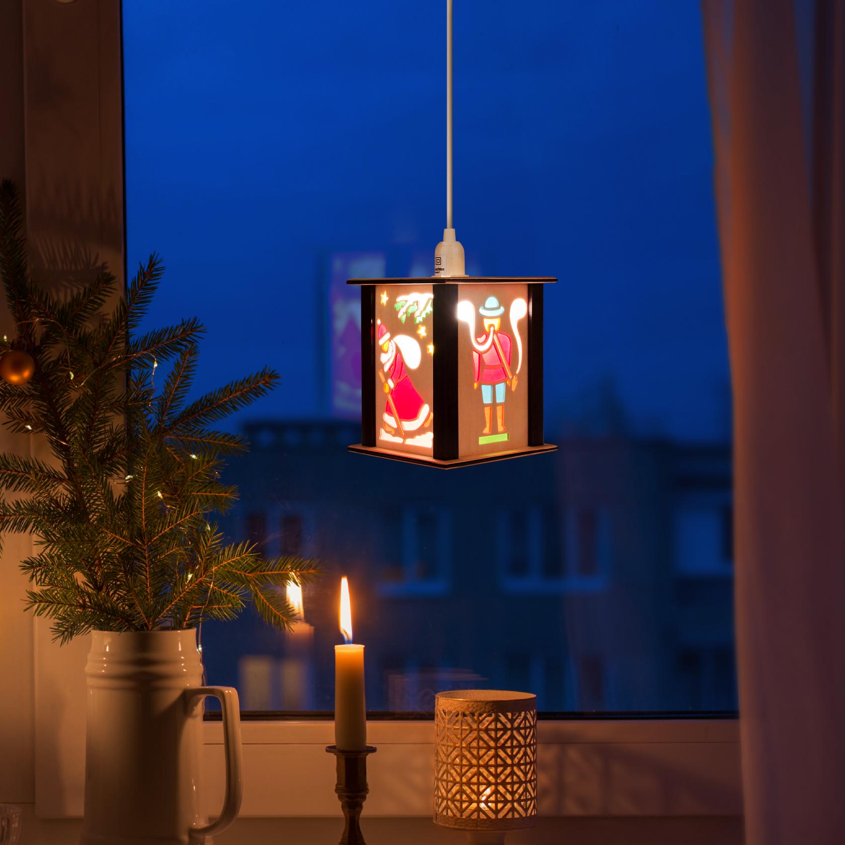 Eine kleine Laterne mit Nussknacker-Motiv hängt beleuchtet vor einem Fenster. Im Vordergrund stehen eine brennende Kerze und ein Zweig.