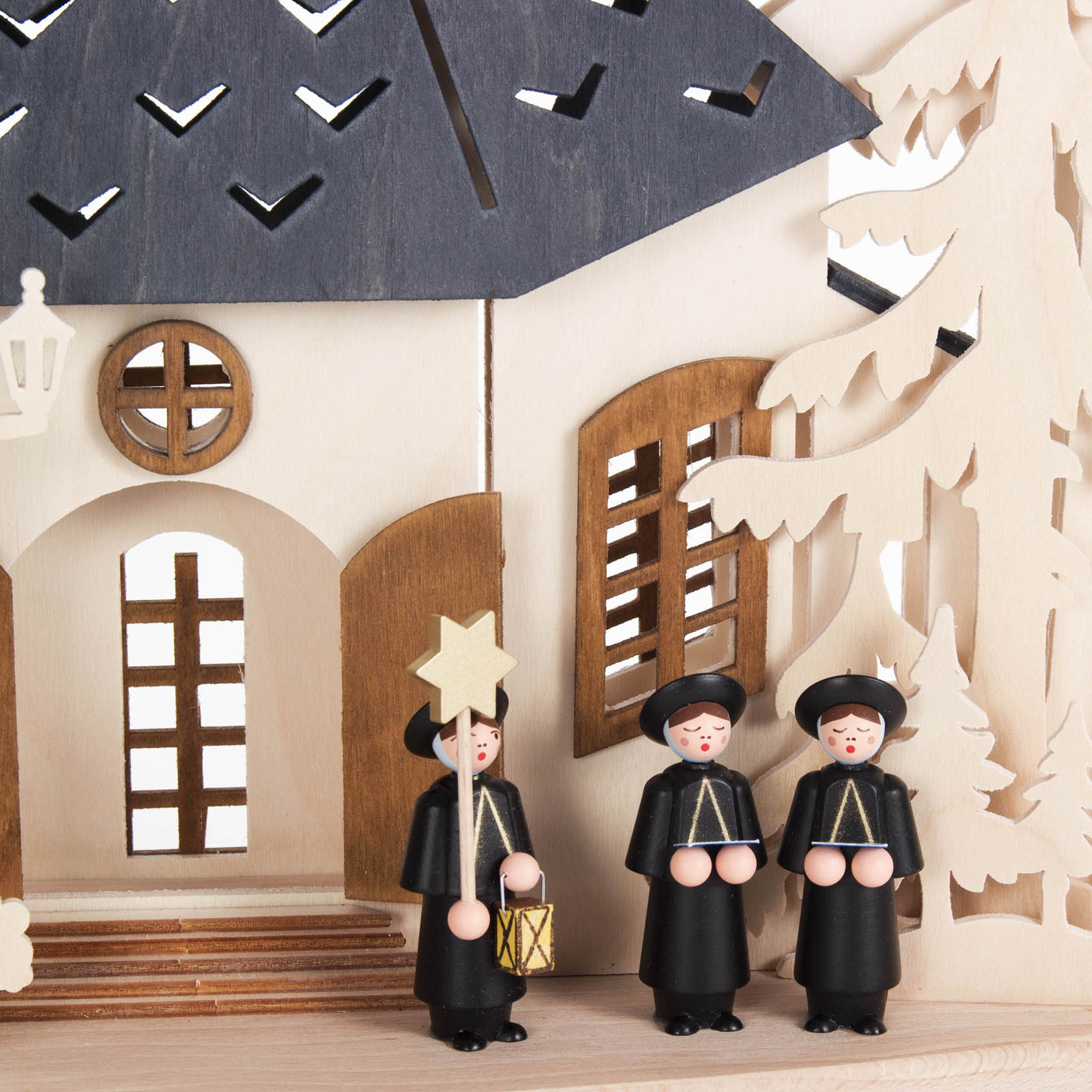 Drei Holzfiguren, die Chorknaben darstellen, mit schwarzen Umhängen und Hüten, stehen vor einem hölzernen Gebäude. Einer trägt einen Stern und eine Laterne.
