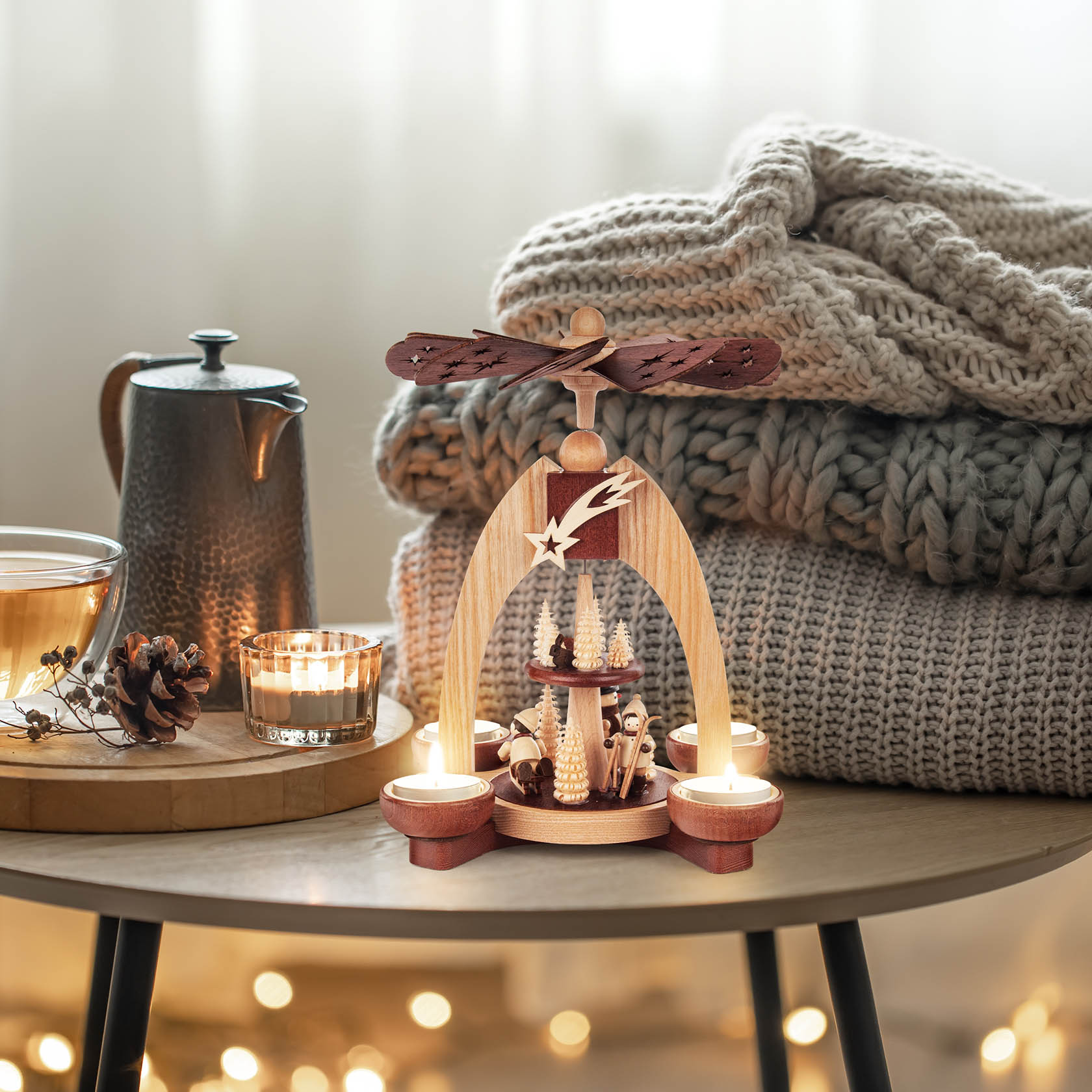 Zweistöckige hölzerne Weihnachtspyramide mit hölzernen Winterfiguren und Teelichthaltern, umgeben von dekorativen Lichtern auf einem Tisch.