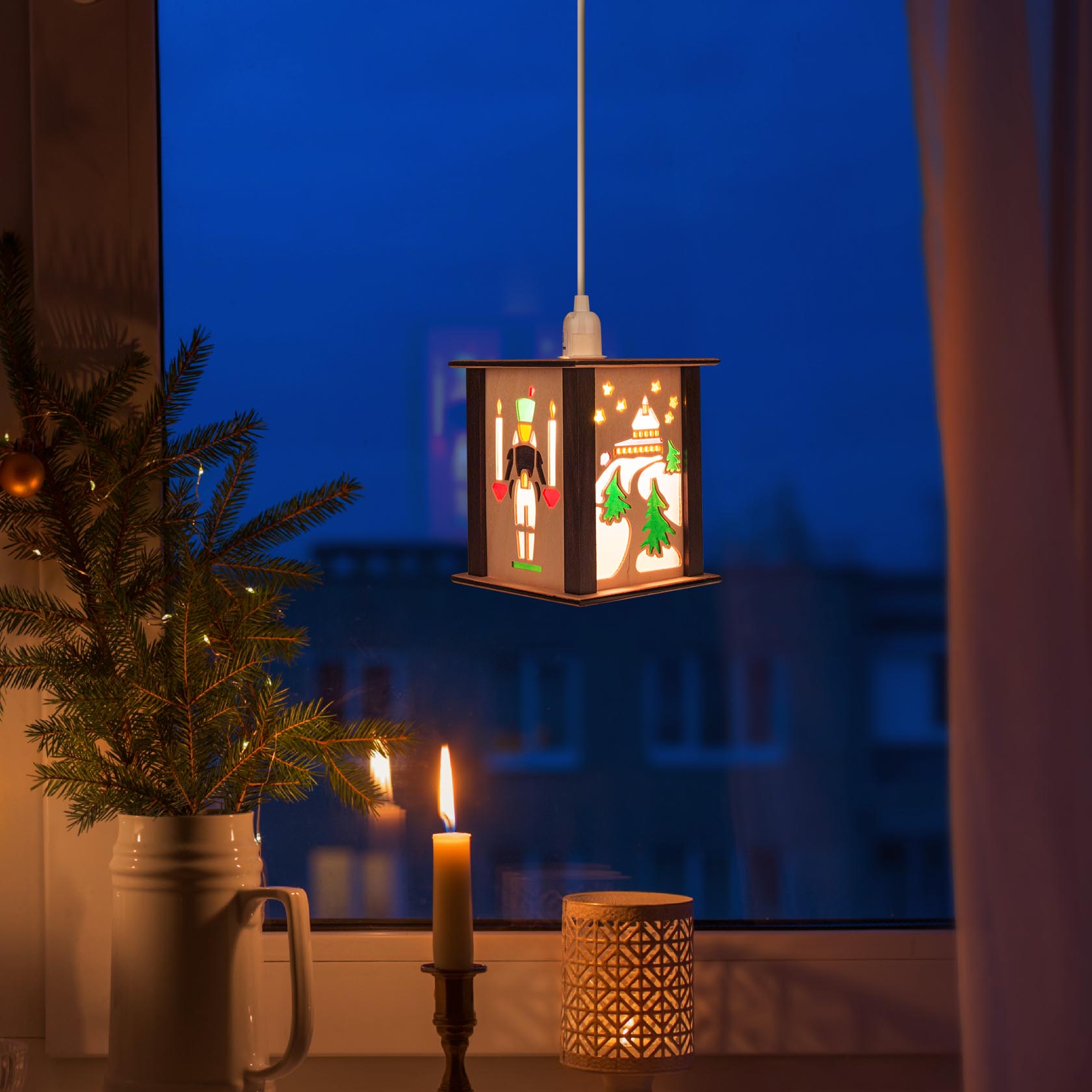 Kleine Laterne mit Motiven der Seiffener Kirche, hängend im Fenster. Beleuchtet, neben einer brennenden Kerze und einem Zweig Tanne auf einem Tisch.