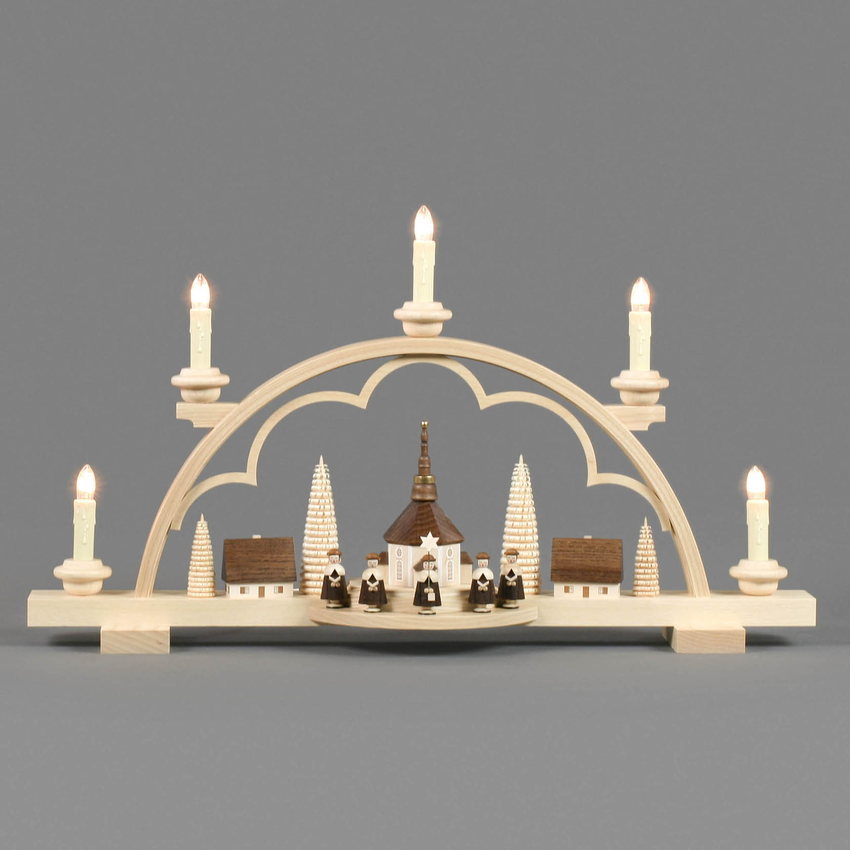 Ein Schwibbogen mit einem Seiffener Dorf und Kurrende, elektrisch beleuchtet. Der Bogen zeigt eine Kirche, flankiert von Kurrende-Sängern und zwei Häusern, mit sechs Kerzen.