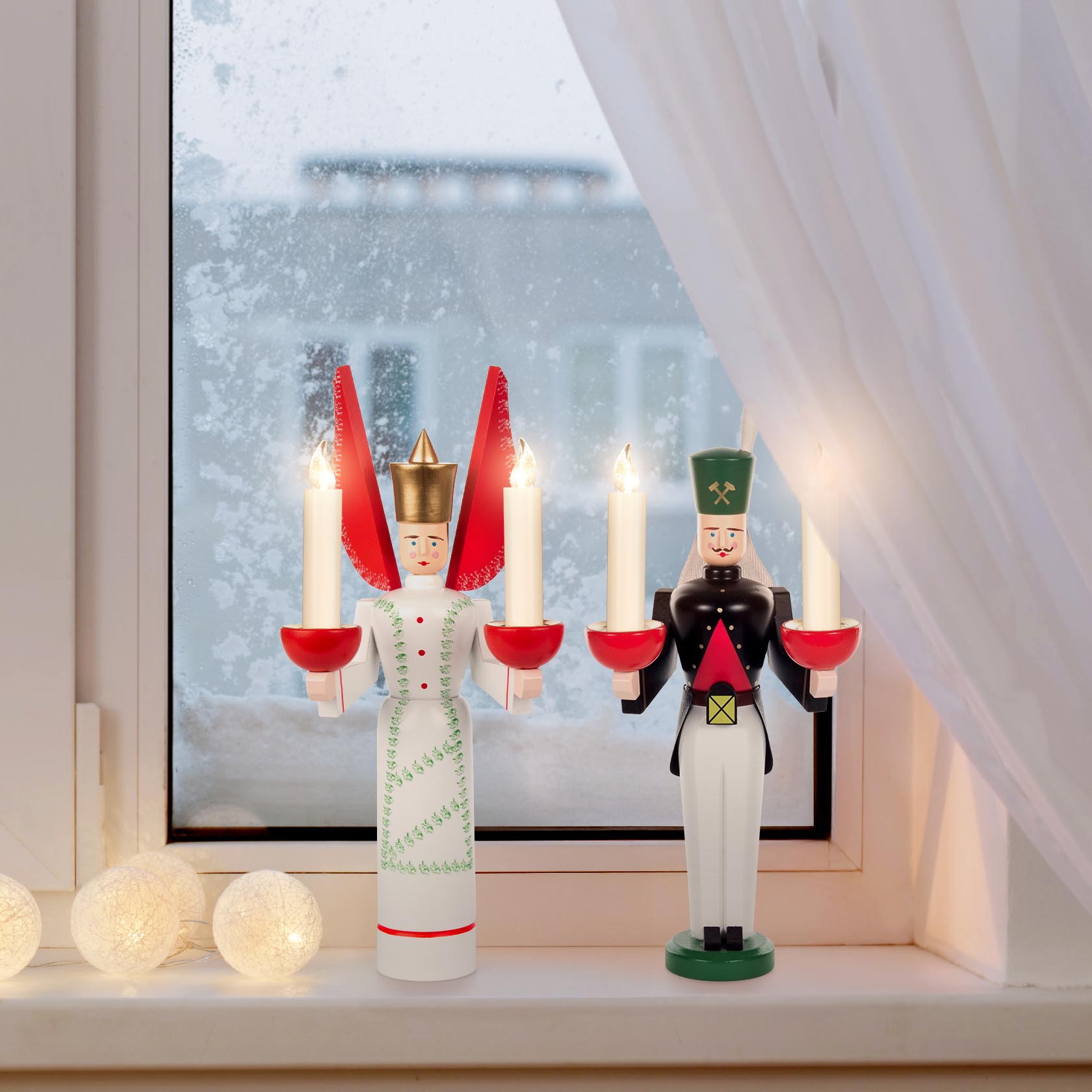 Holzfiguren von Engel und Bergmann, beide je zwei Kerzen haltend, stehen auf einer Fensterbank. Der Engel trägt eine getupfte Schürze.