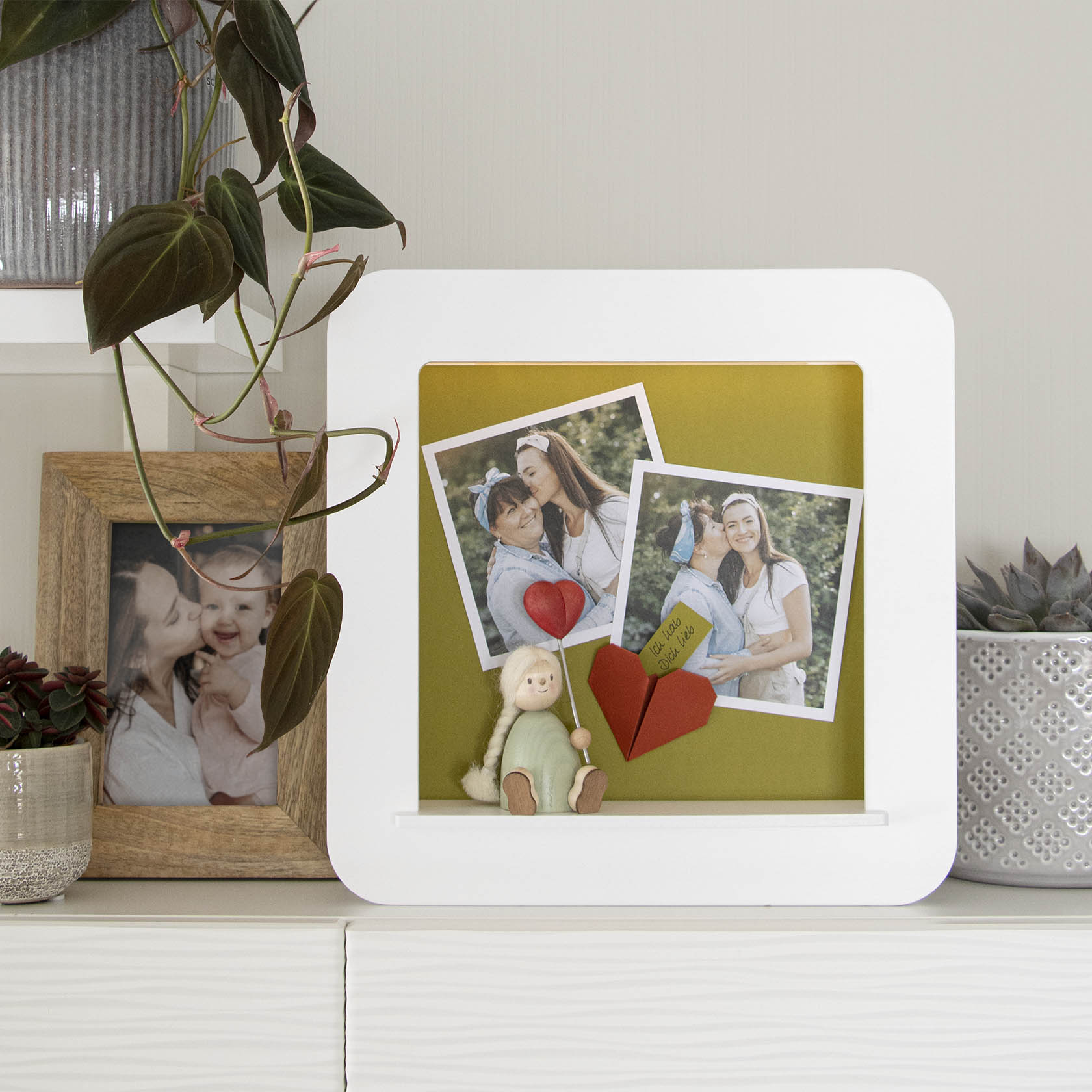 Weißer Objektrahmen mit LED-Beleuchtung, mit zwei Polaroid-Fotos, einer kleinen Puppe und Origami-Herz als Dekoration.