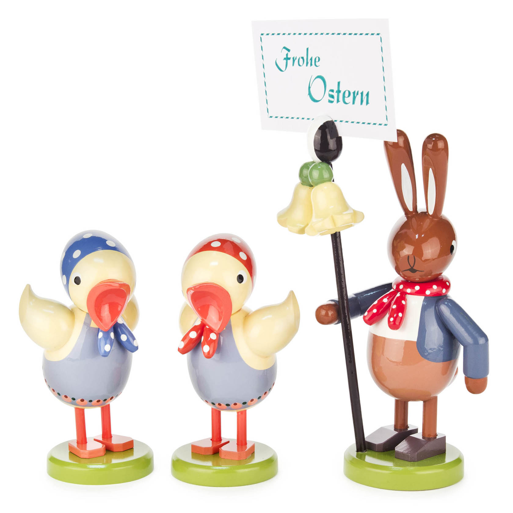 Zwei gelbe, bemalte Holzenten mit blauen Hüten neben einem Hasen, der ein Schild mit 'Frohe Ostern' hält, alle auf grünem Boden stehend. im Dregeno Online Shop günstig kaufen