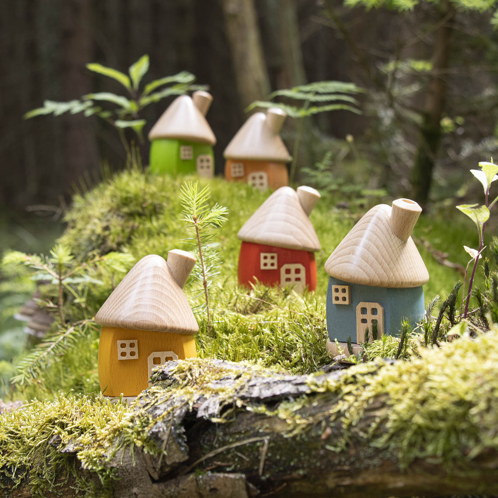 Vier kleine Holz-Räucherhäuser mit konischen Dächern in den Farben Gelb, Rot, Grün und Blau stehen in einem moosigen Waldgebiet.