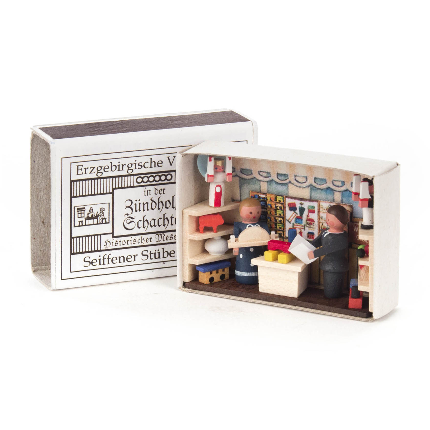 Miniatur-Zündholzschachtel zeigt einen traditionell gestalteten Messestand mit zwei Holzfiguren. Eine Person überreicht einem Kind ein Geschenk.