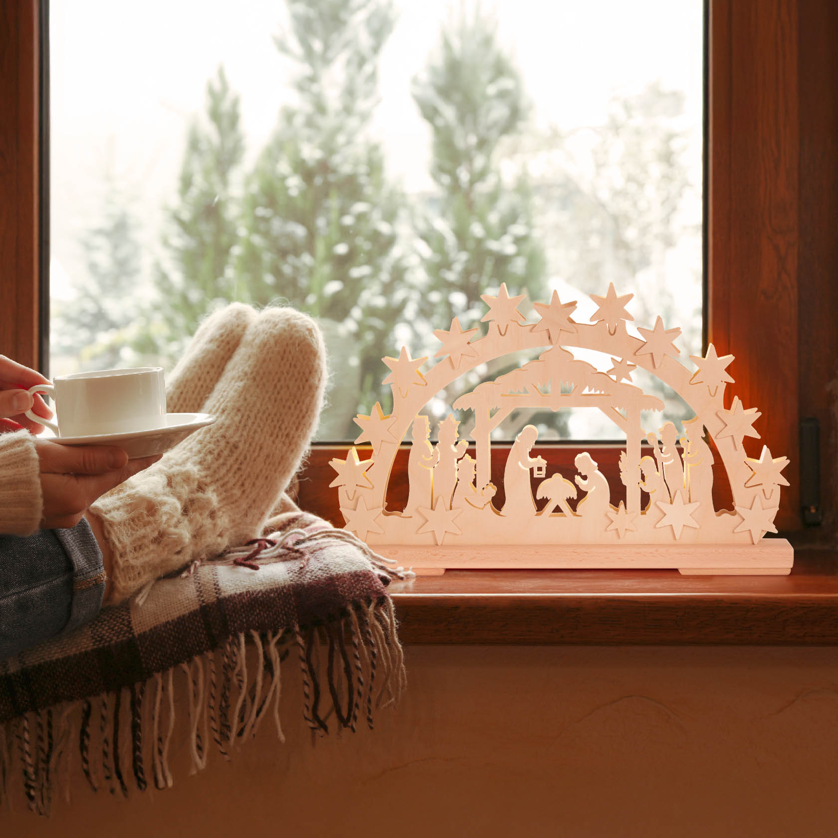 A small, electrically illuminated arch (Schwibbogen) featuring the Nativity scene, placed on a window sill with a person sitting nearby, cozy with a cup and woolen socks.