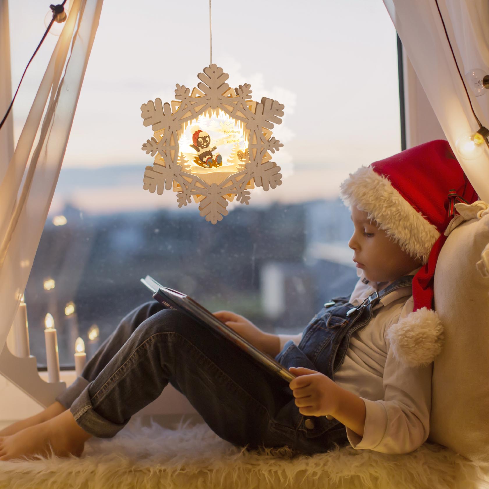 Ein Kind mit Weihnachtsmütze sitzt am Fenster, liest ein Buch und neben ihm hängt ein leuchtendes, sternförmiges Fensterbild.