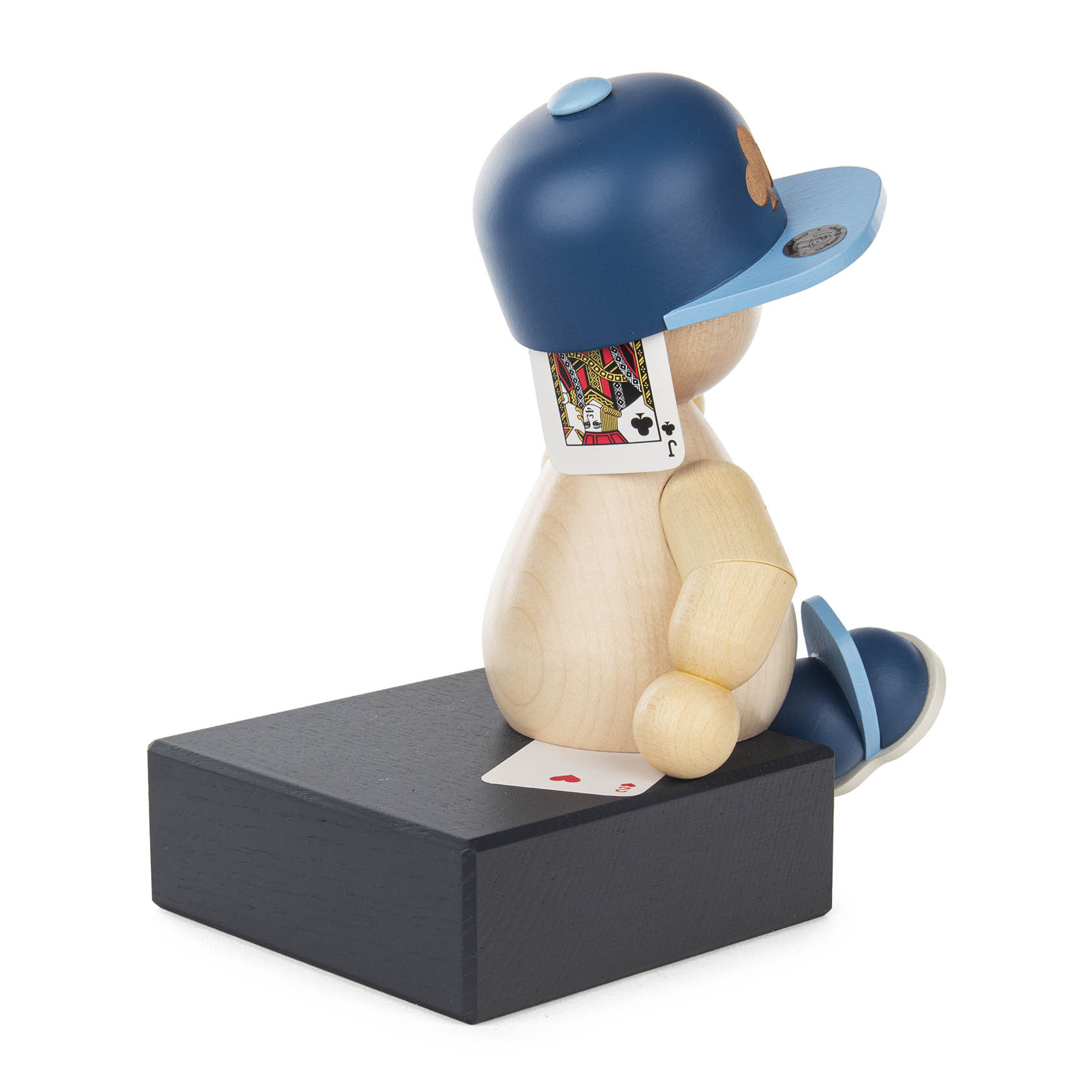 Holzpuppe in Baseballkappe sitzt auf schwarzem Sockel mit Spielkarte am Rücken.