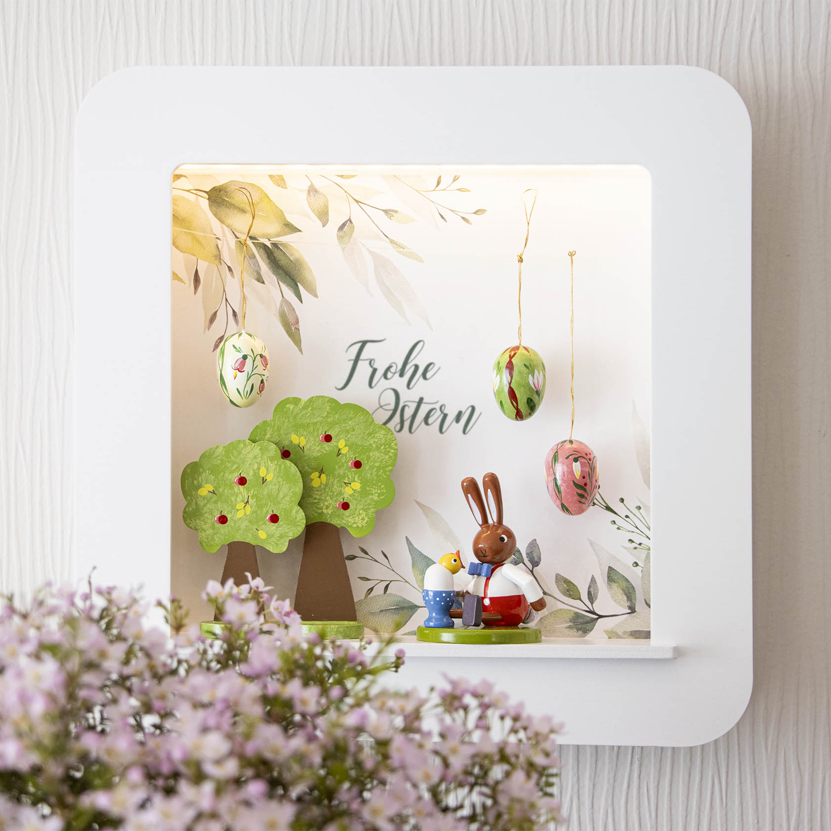 Beleuchteter Objektrahmen mit Osterdeko: Hase mit Küken, gemusterte Ostereier und Text 'Frohe Ostern'. Blumen im Vordergrund.