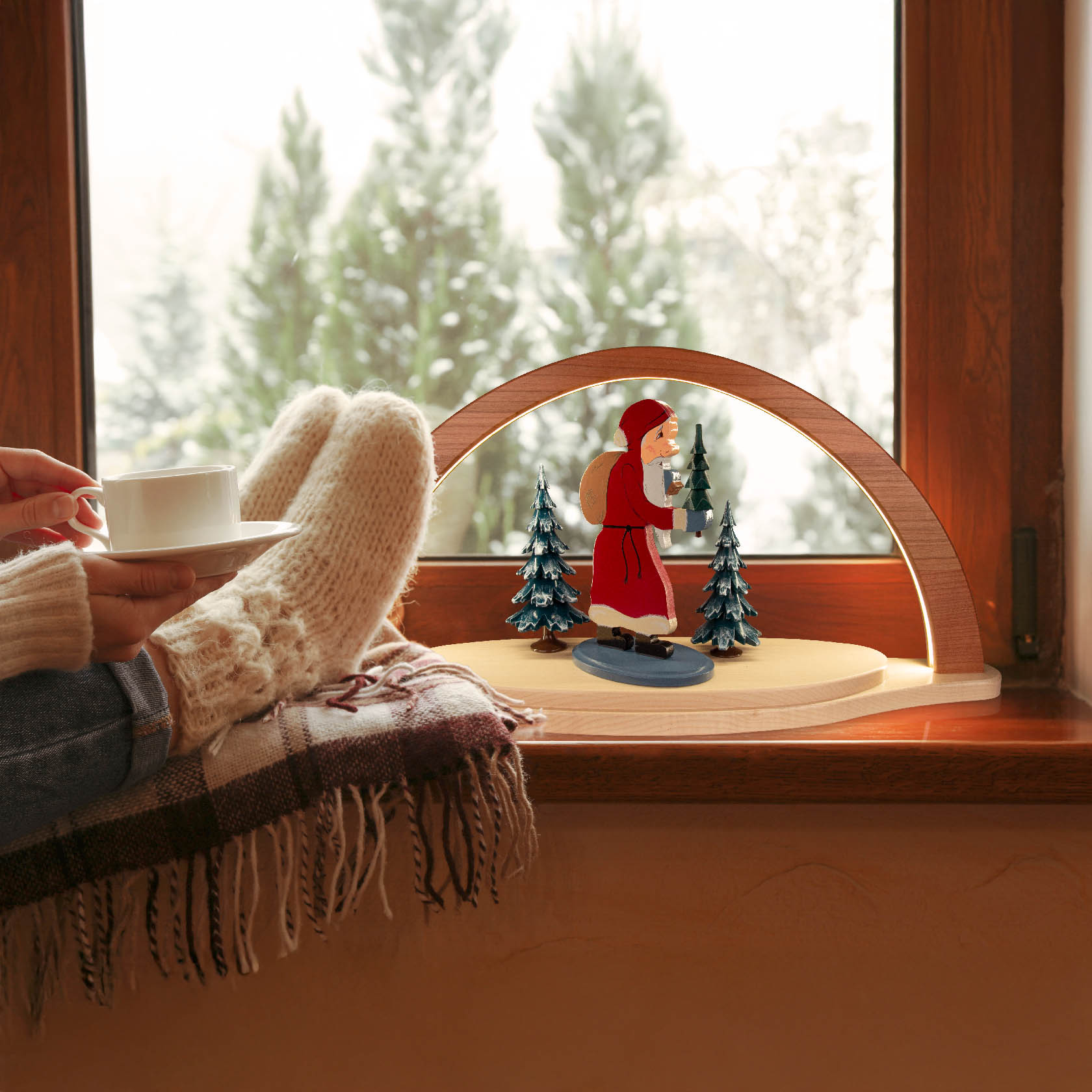 Sitzende Person mit Wollsocken hält Tasse und blickt auf beleuchteten Holz-Schwibbogen mit einer rot gekleideten Figur und Tannen auf dem Fensterbrett.