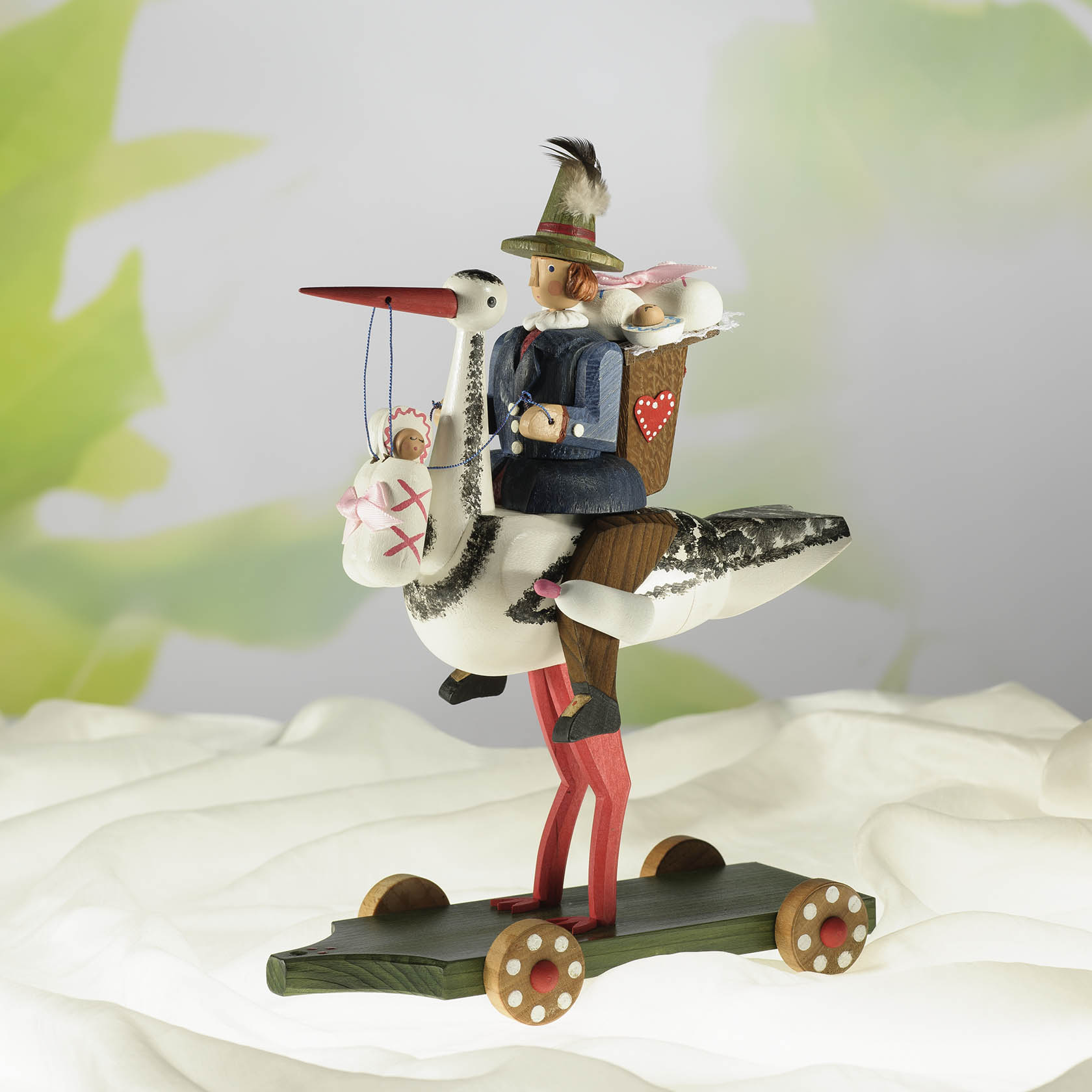 Erzgebirgische Holzfigur eines stolzen Storches mit Zylinder, auf dem ein Reiter in Tracht sitzt. Hinter ihm ein Korb mit Stoffpüppchen.