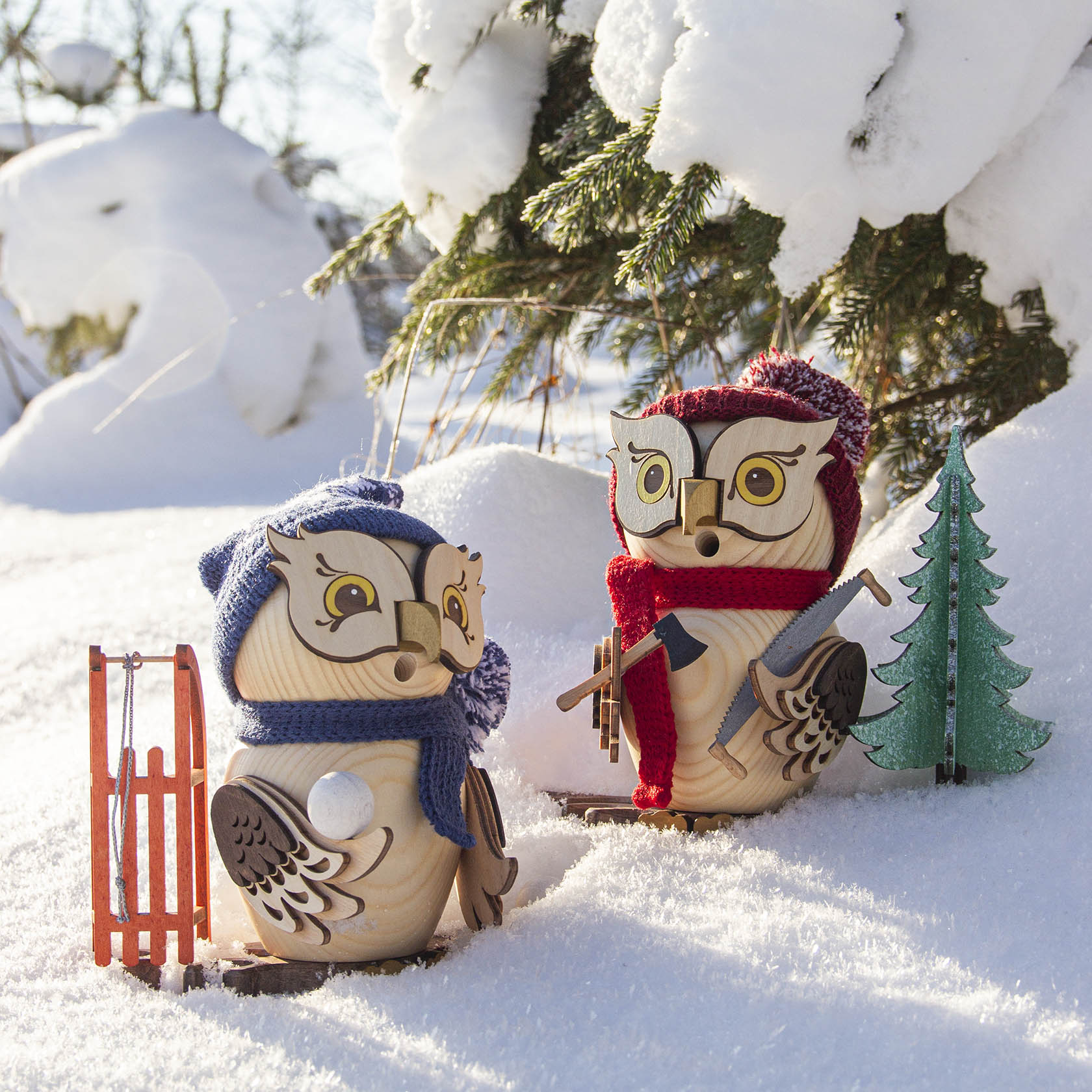 Zwei hölzerne Räuchereulen stehen im Schnee. Eine trägt einen blauen Schal und hält einen Schlitten. Die andere hat einen roten Schal und eine Spielzeug-Axt.