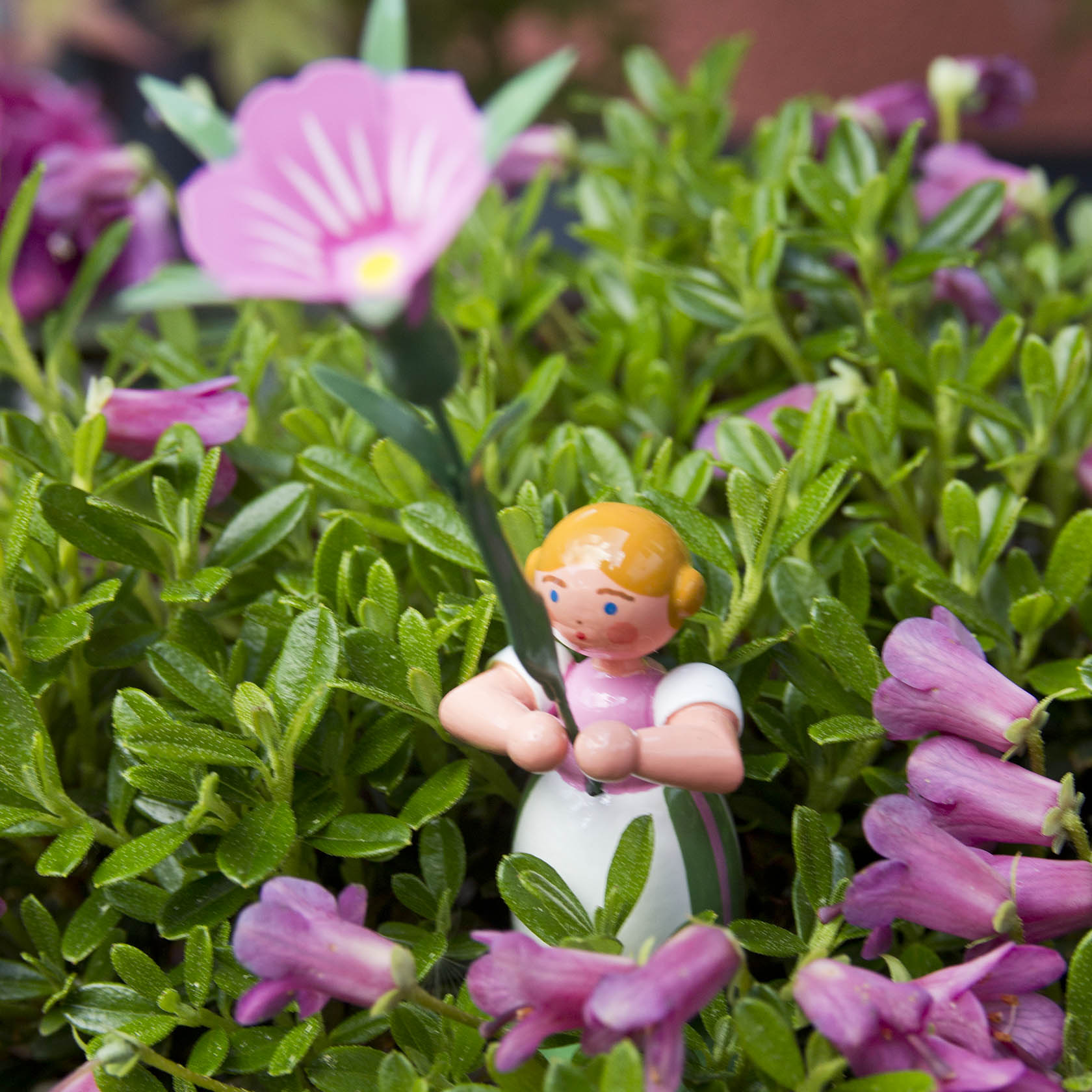 Eine kleine Figur eines Mädchens mit blonden Haaren hält eine rosa Winde inmitten von lila-rosa Blumen und grünem Laub.