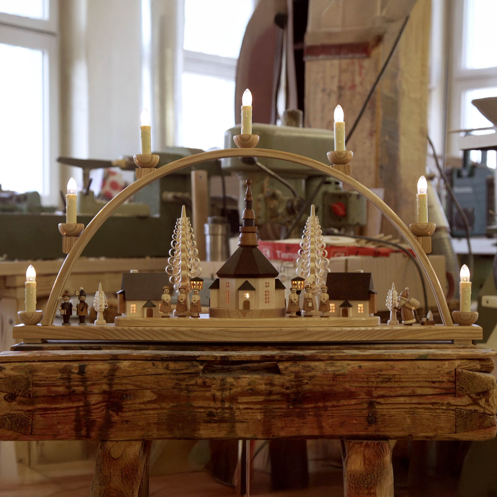 Ein Schwibbogen zeigt ein Seiffener Dorf mit elektrisch beleuchteten Gebäuden und Laternen. Im Zentrum steht eine Kirche, umrahmt von Bäumen und Figuren.