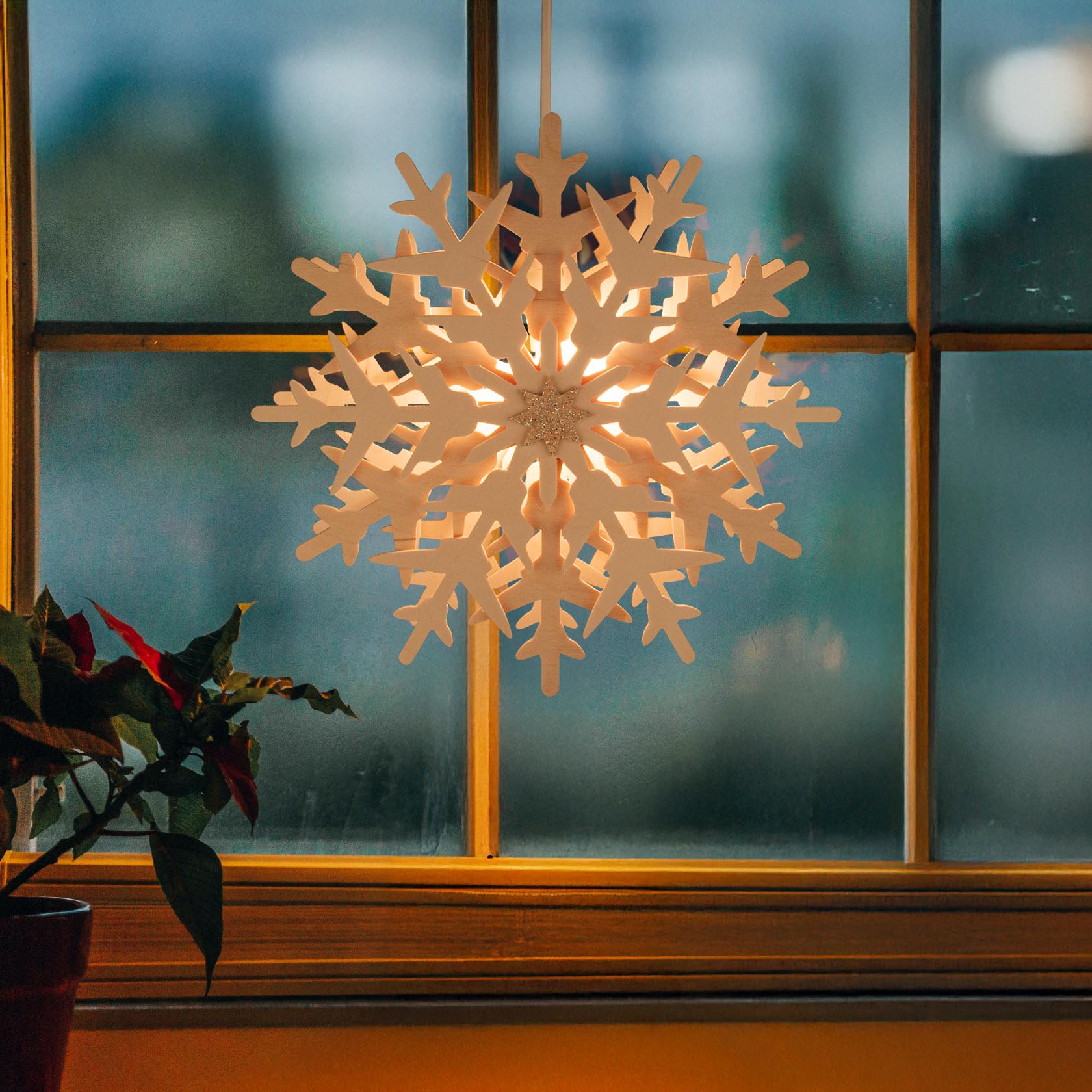 Illuminated snowflake decoration hanging in a window, with a soft glowing light from within and a potted plant nearby.