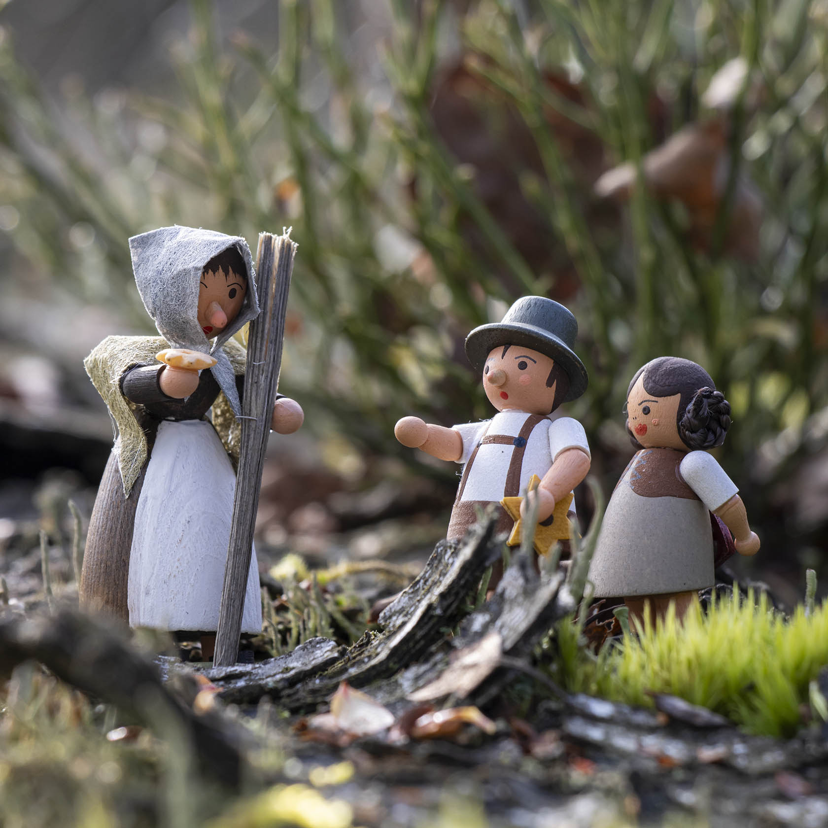 Eine Puppenhexe mit Kapuze und Umhang kniet vor zwei Puppenbauernkindern im Freien. Die Kinder tragen traditionelle Kleidung und schauen die Hexe an.