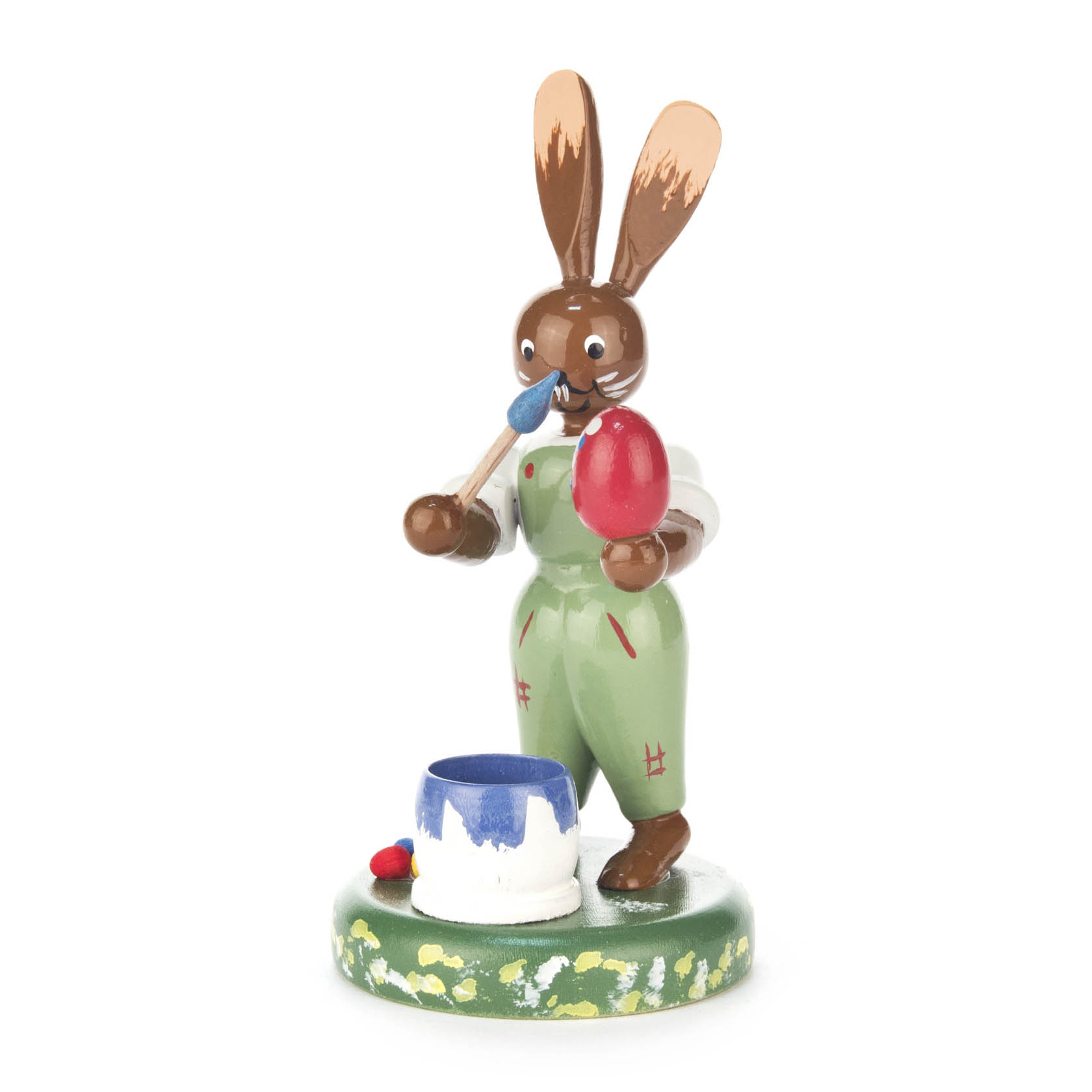 Holzfigur eines Hasen in grüner Latzhose, der ein rotes Ei mit einem blauen Pinsel bemalt. Vor ihm ein blauer Farbtopf. im Dregeno Online Shop günstig kaufen
