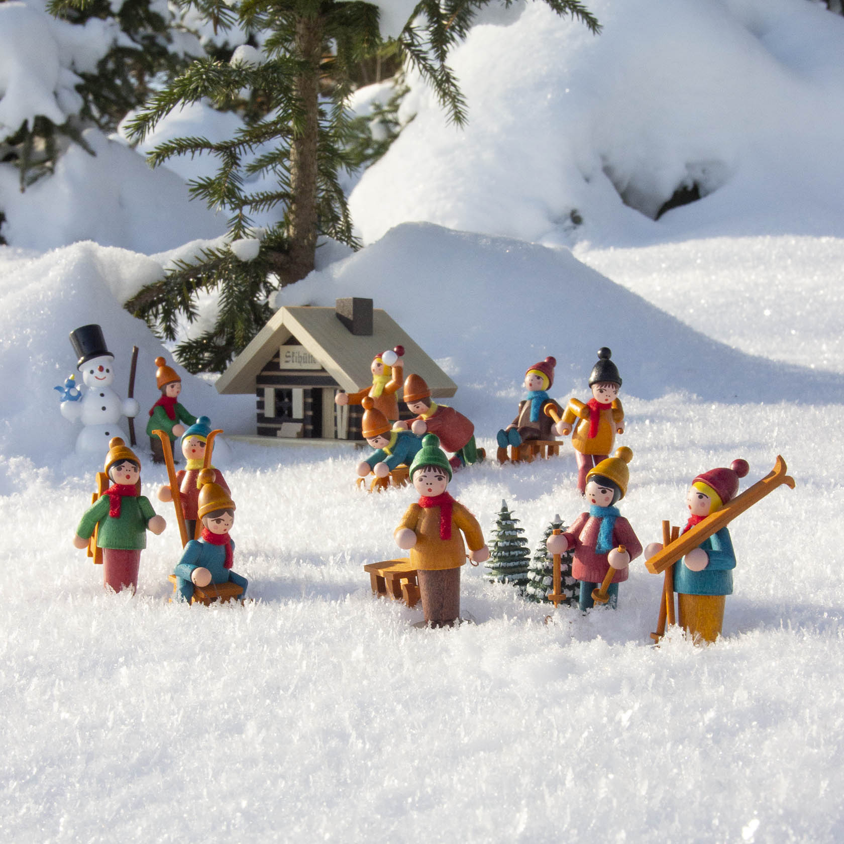 Eine verschneite Skiszene mit farbenfrohen Holzfiguren, die Skifahren oder Schlittenfahren. Im Hintergrund steht eine kleine Holz-Räucherhütte.