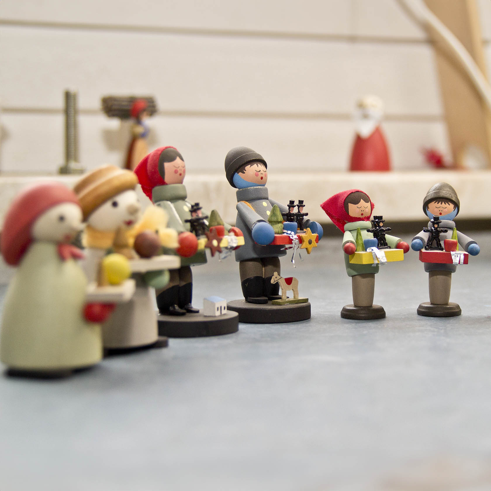 Holzfiguren von Kindern, zwei tragen Tabletts mit Stollen und Kerzen, eine Gruppe mit roten und dunkelgrauen Mützen in einer Werkstatt.