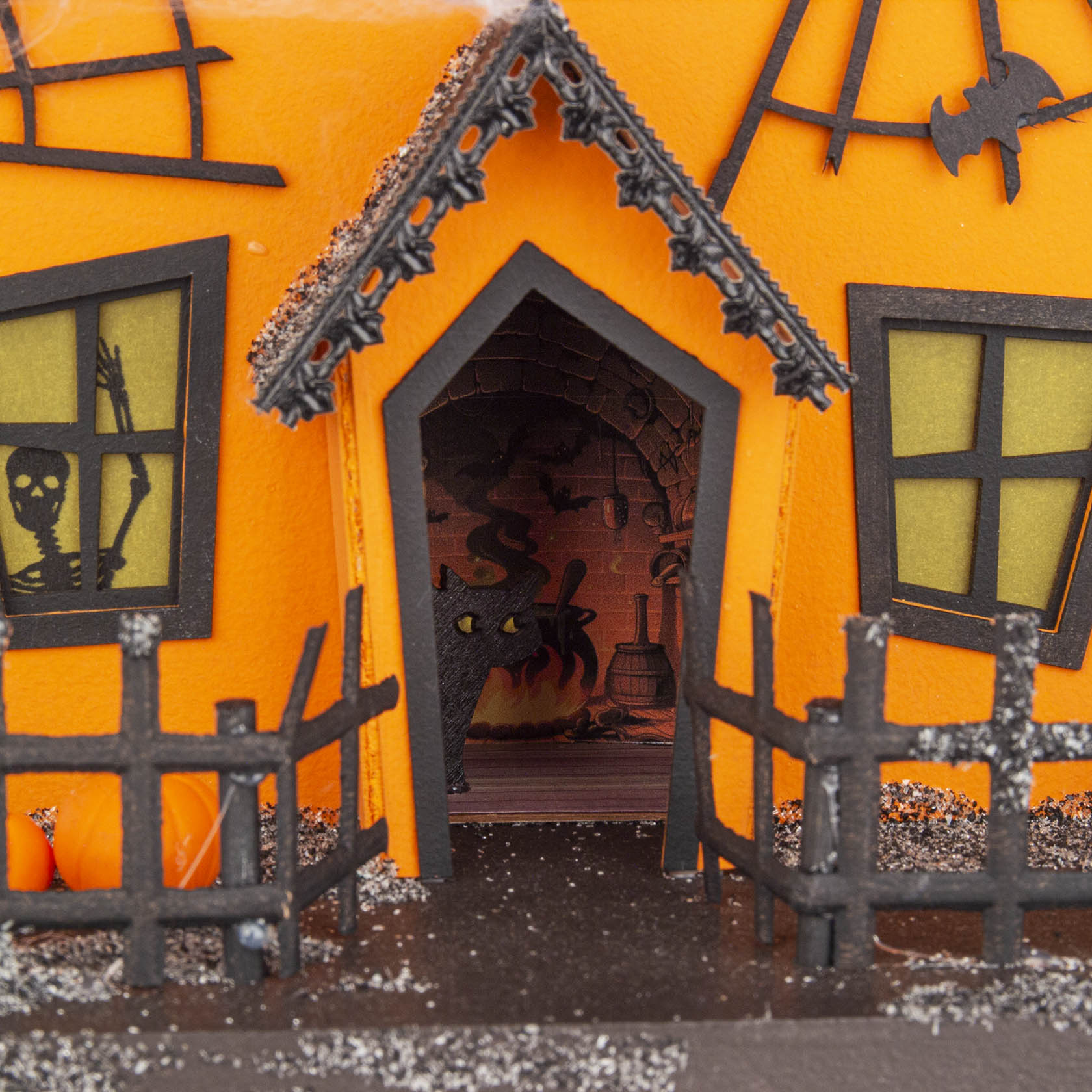 Ein dekoratives Kürbishaus mit schiefen schwarzen Holzfenstern und Türrahmen. Eine Skelettfigur sowie ein Hexenkessel sind im Inneren sichtbar.