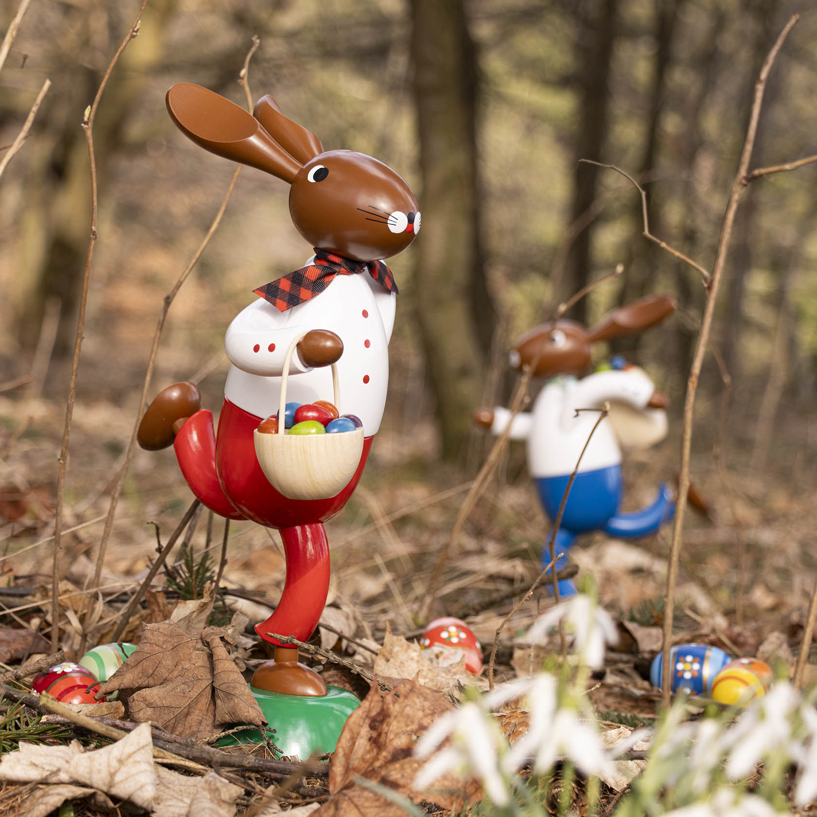 Zwei hasenförmige Figuren im Wald, jede mit Ostereiern. Vorderer Hase trägt einen roten Anzug und hat einen Korb, hinterer Hase in Blau mit einer Kiepe.