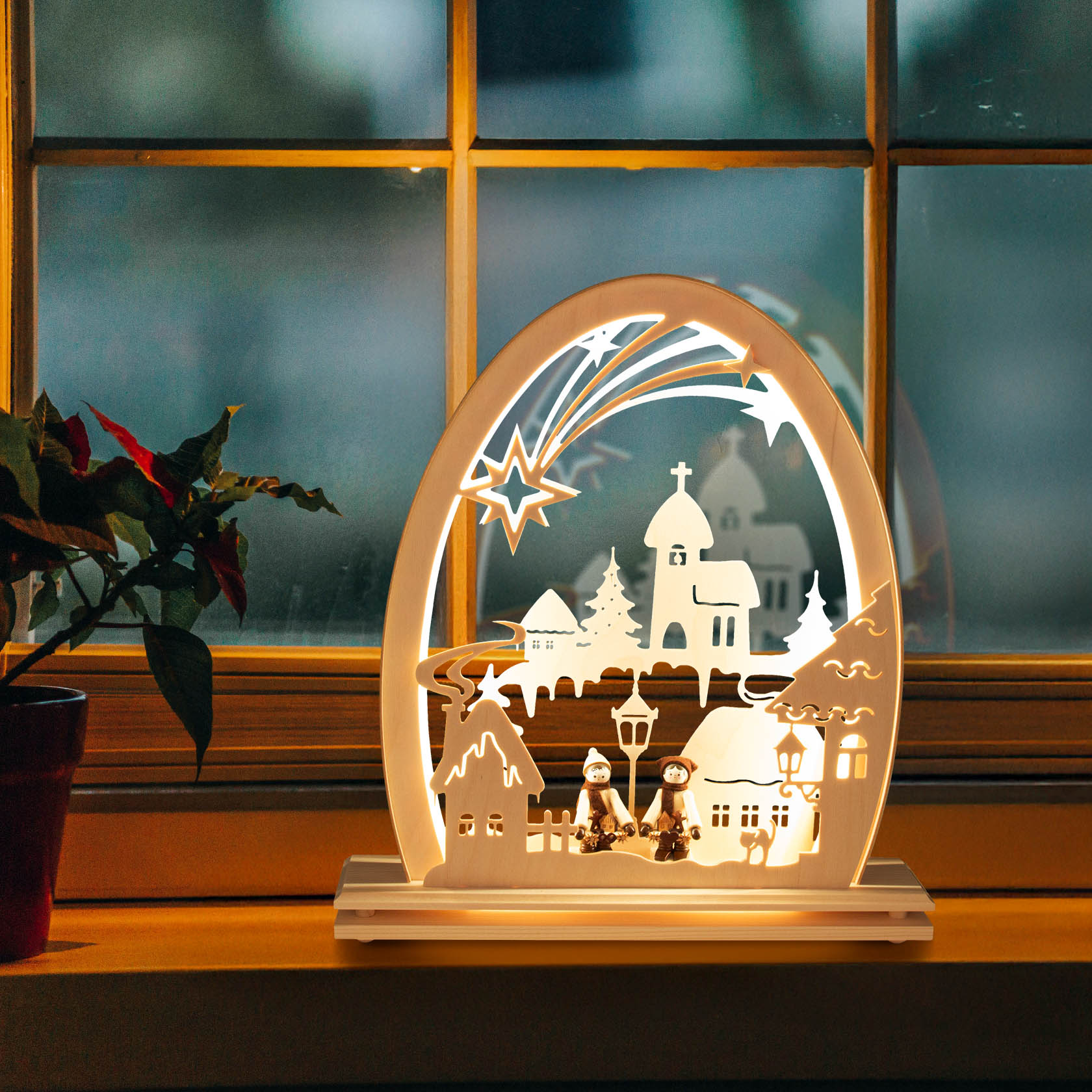 Ein beleuchteter Erzgebirgsbogen auf einer Fensterbank zeigt ein winterliches Dorf mit LED-Beleuchtung, zwei Figuren sitzen unter Straßenlaternen.