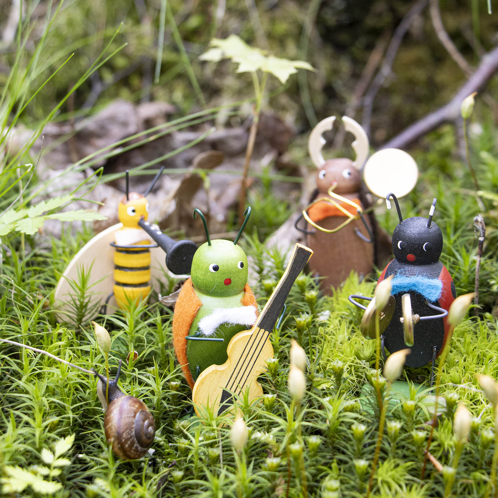 Sechs bunte Insektenfiguren im Moos mit Musikinstrumenten, darunter eine grüne Figur mit Gitarre und eine schwarze mit Saxophon, ebenfalls eine Schnecke.