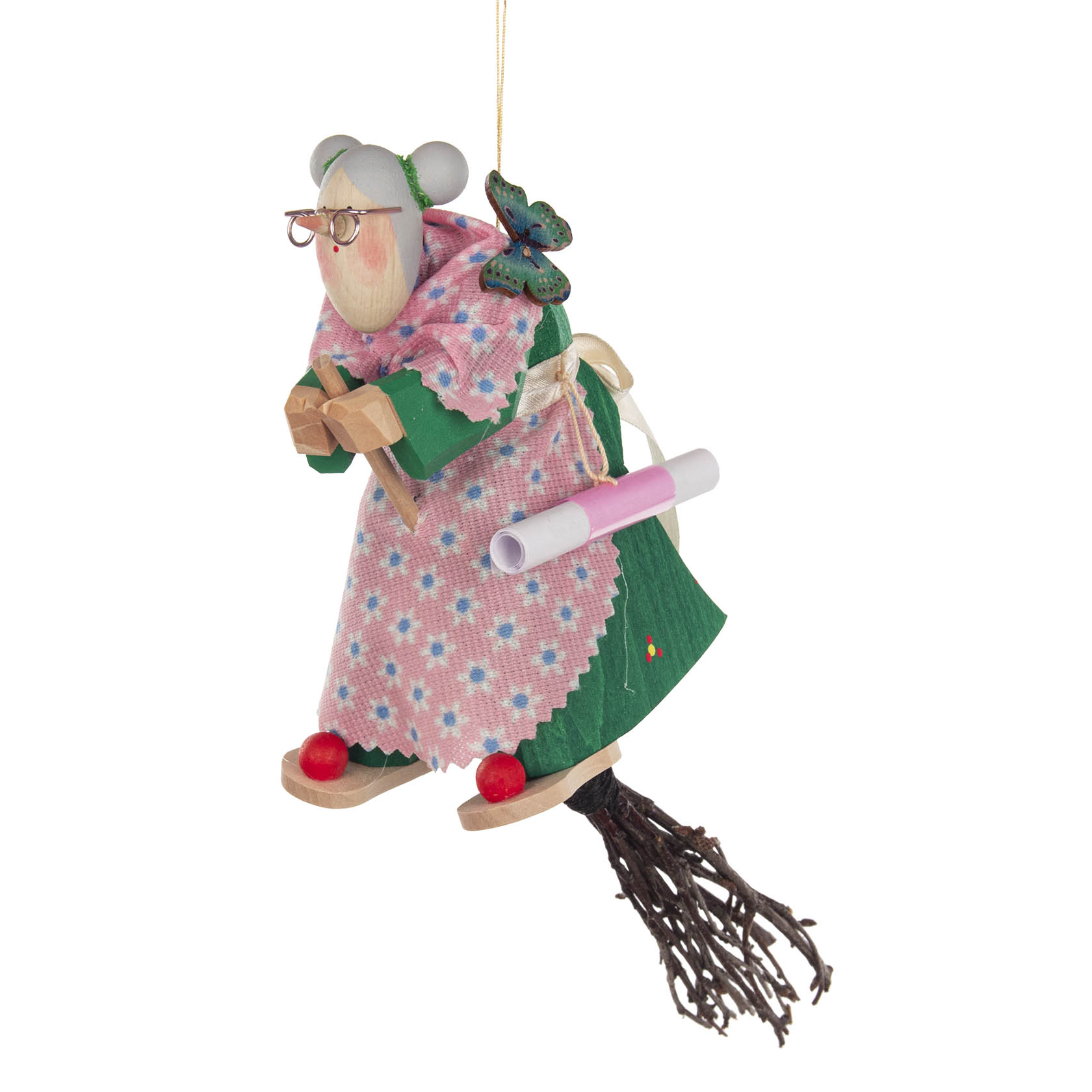 A wooden witch figurine flying on a broomstick, wearing glasses, a green dress, and a pink floral shawl with a butterfly decoration on her shoulder.