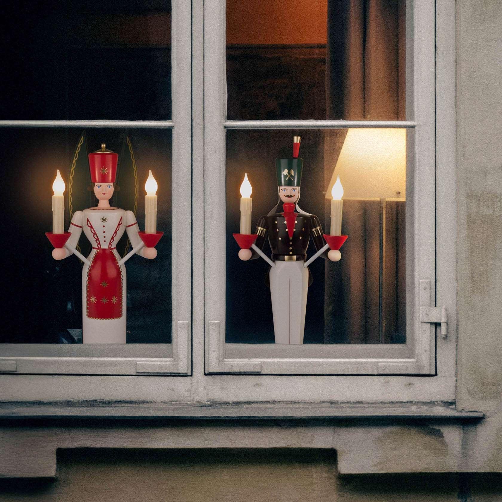 Ein Lichterbergmann und eine Lichterfrau stehen nebeneinander vor einem Fenster. Beide halten elektrische Kerzenhalter mit jeweils zwei Lichtern.