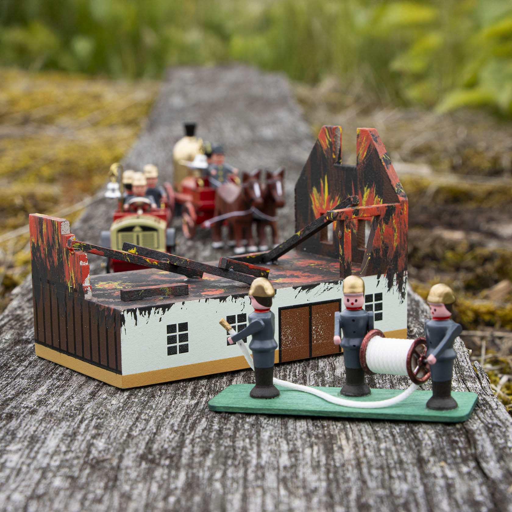 Model of a burning house with three firefighter figurines, one holding a hose, and a horse-drawn fire brigade approaching.