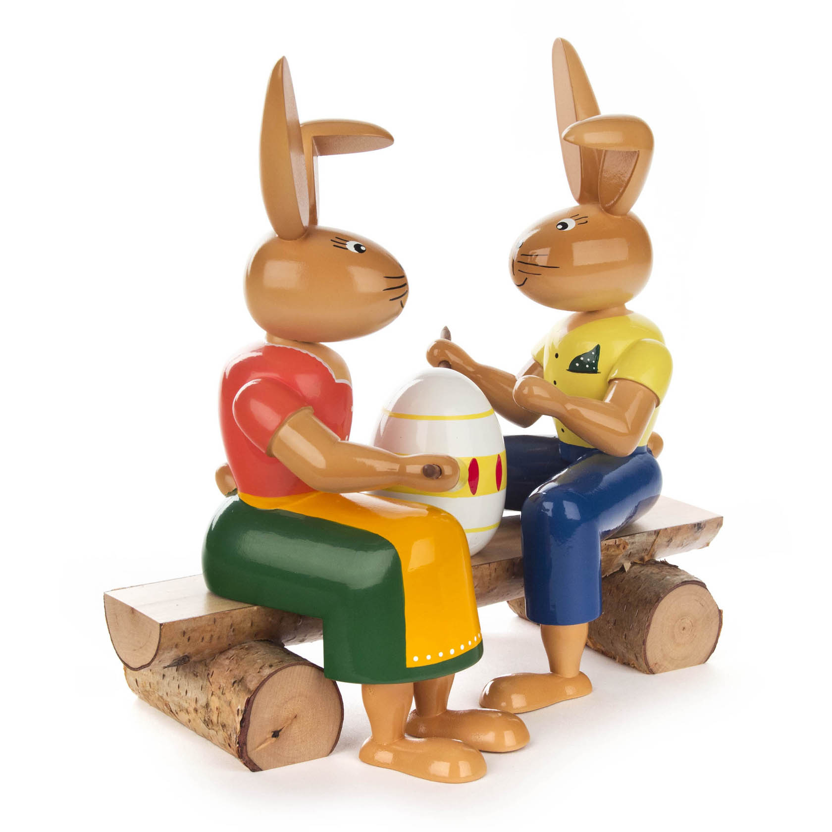 Zwei handgefertigte Holzhasen in bunten Kleidern sitzen auf einer Bank aus Holzscheiten und bemalen gemeinsam ein Osterei.