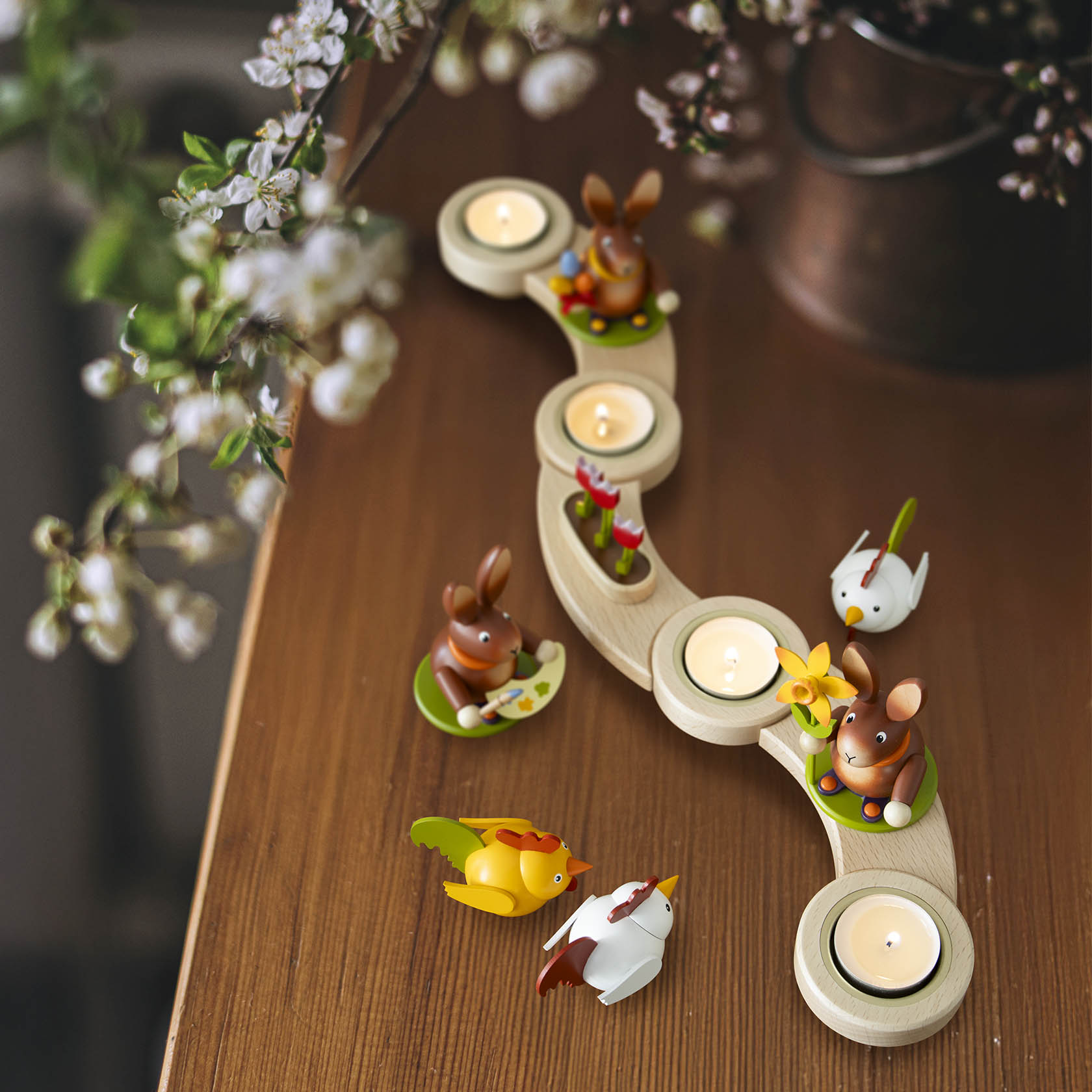 Drei Teelichter in einem geschwungenen Halter aus Holz, dekoriert mit bunten Tierfiguren und einer roten Tulpenblüte.