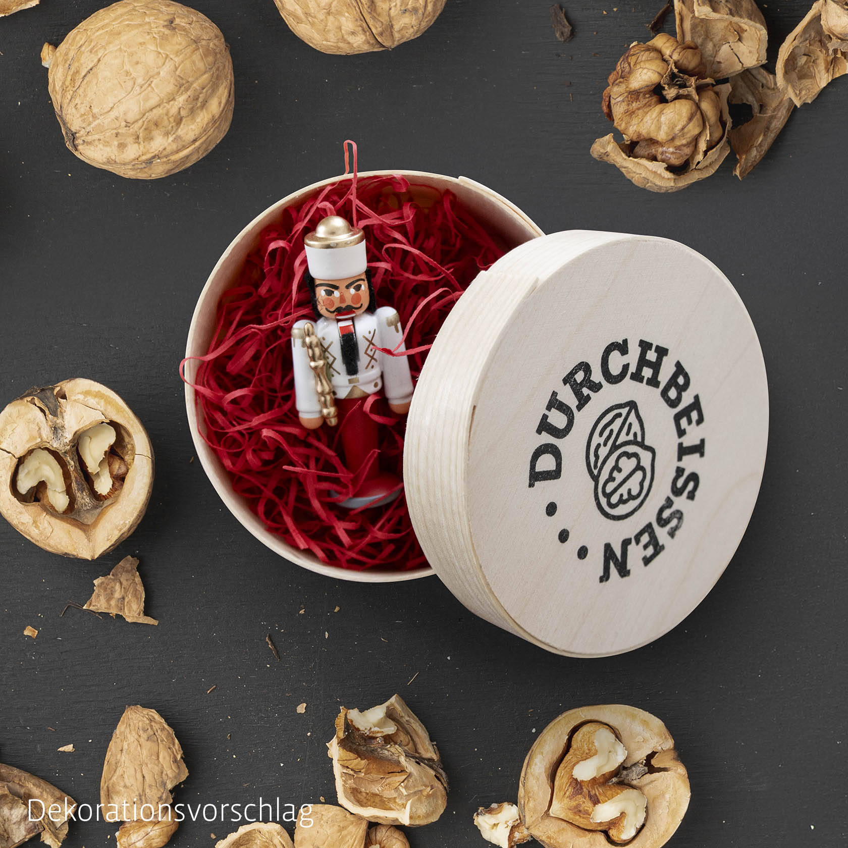 Ein Mini-Nussknacker in einer runden Spandose mit rotem Füllmaterial. Die Dose hat den Schriftzug 'DURCHBEISSEN' mit Nussmotiv auf dem Deckel. im Dregeno Online Shop günstig kaufen