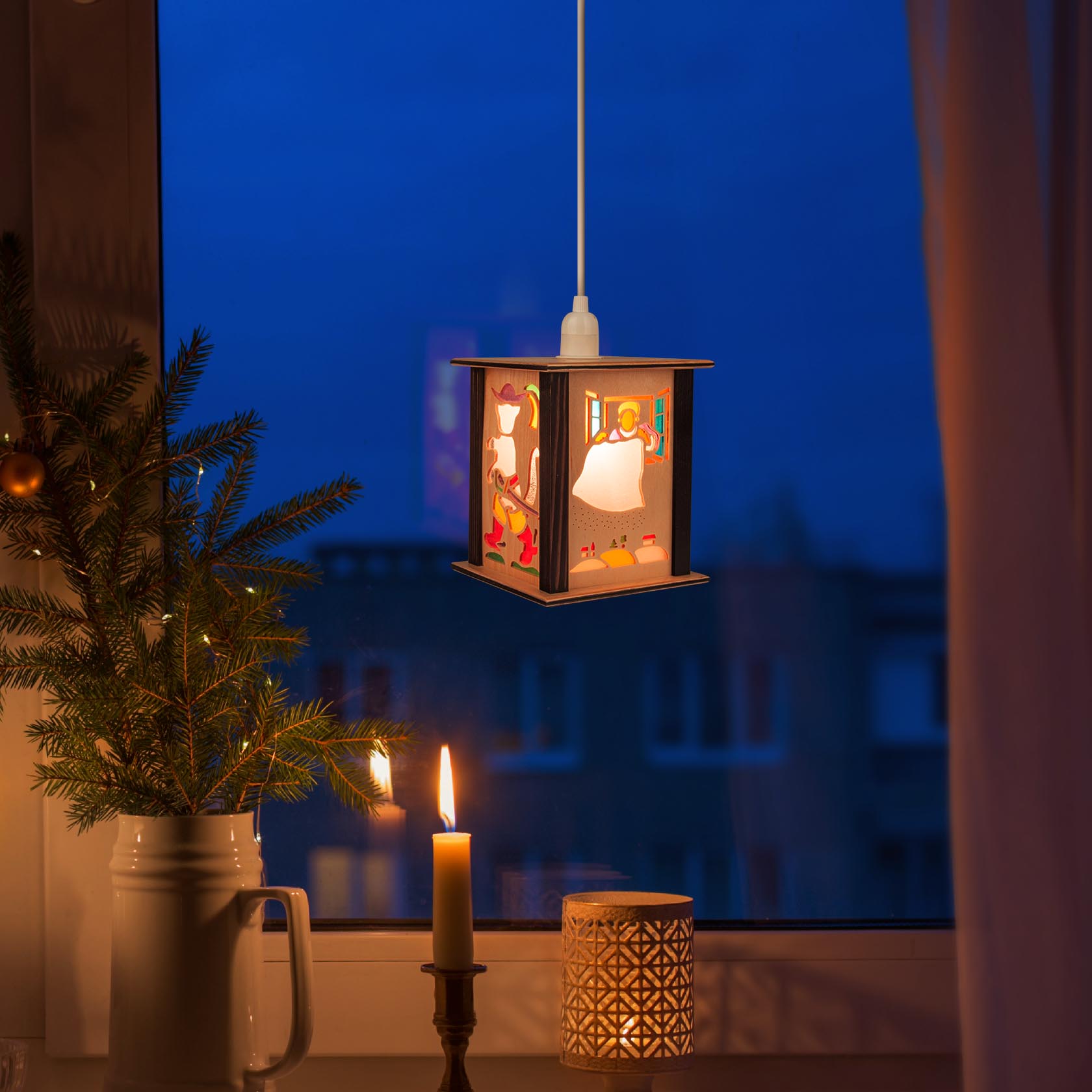 Eine dekorative Hängelaterne zeigt Märchenszenen in einem Fenster. Daneben ein kleiner Weihnachtsbaum und eine brennende Kerze.