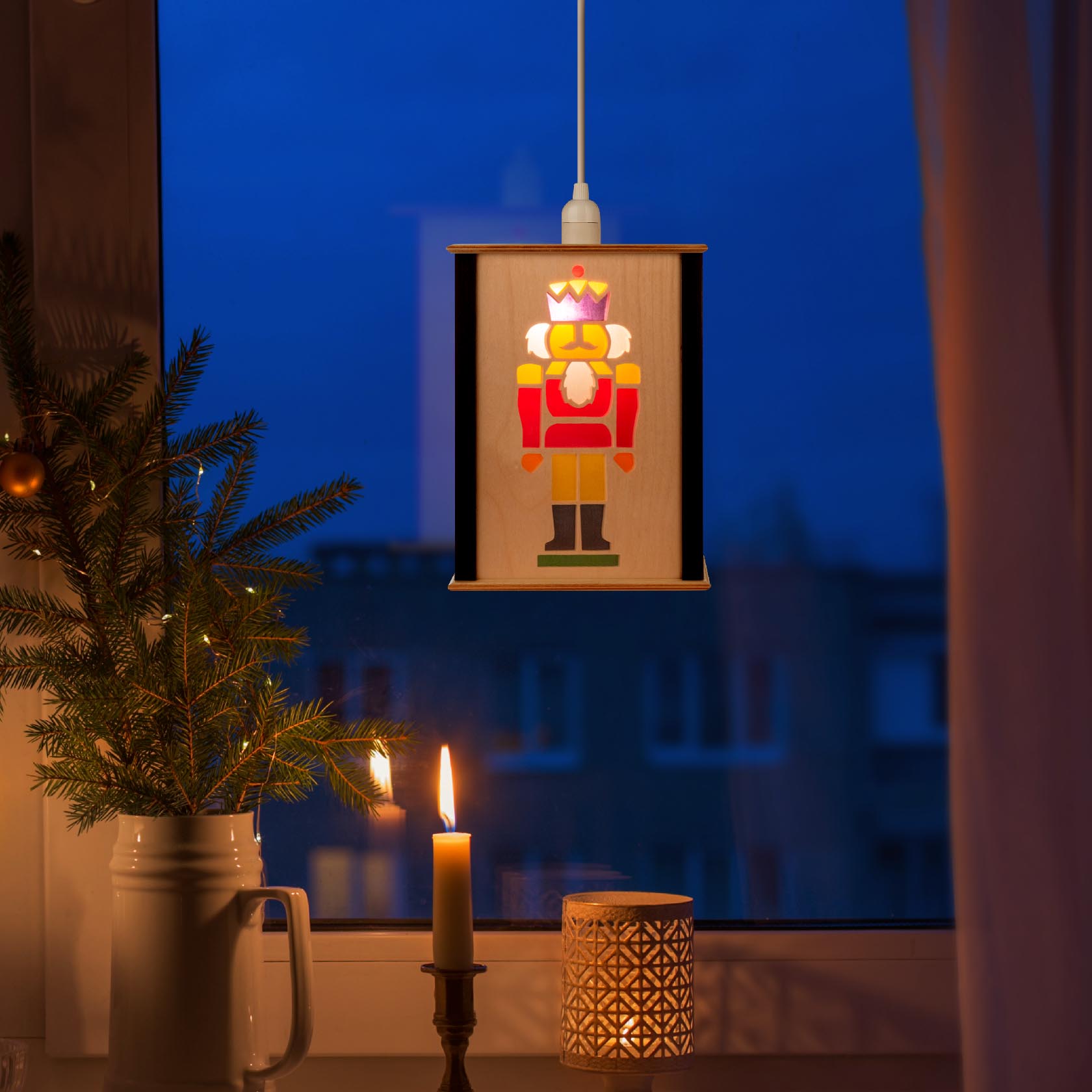 Eine erleuchtete Fensterlaterne mit einem Nussknackerdesign hängt vor einem Fenster. Auf der Fensterbank stehen eine brennende Kerze und ein Tannenzweig.